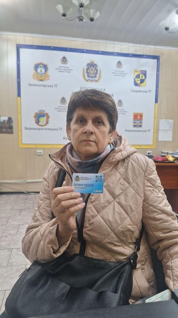На світлині у напіввідкритий зріст зображено жінку старшого віку з числа внутрішньо переміщених осіб з Херсонщини під час отримання допомоги в Одеському координаційному центрі «Вільні разом». Вона сидить у кімнаті та тримає у правій руці перед собою пластикову «Картку Херсонщини» синьо-блакитного кольору з логотипом проєкту «Вільні разом Херсонщина» та QR-кодом. Жінка з короткою темною зачіскою одягнена в бежеву стьобану осінню куртку з капюшоном та теплий шарф, а на плечі у неї висить велика чорна шкіряна сумка. Позаду жінки на світлій стіні кабінету закріплено великий офіційний білий стенд із гербами та написами територіальних громад області, серед яких чітко видно логотипи «Зеленопідська ТГ», «Скадовська ТГ», «Верхньорогачицька ТГ» та «Новокаховська ТГ».