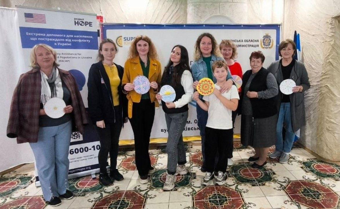 На світлині зображено групу переселенців із Херсонщини, серед яких є жінки, дівчата та один хлопчик, після завершення арттерапевтичних занять в Одеському координаційному центрі «Вільні разом». Учасники заходу вишикувалися в один ряд у приміщенні з візерунчастою кахельною підлогою та тримають у руках власноруч створені кольорові ресурсні мандали й круглі малюнки-обереги. Присутні одягнені у повсякденний осінній одяг: джинси, куртки, светри, кофти та кардигани, а одна з дівчат у центрі виділяється яскравим гірчичним жакетом. За спинами учасників встановлено великий горизонтальний синій банер із логотипом «SUPPORT KHERSON» та офіційним гербом Херсонської обласної військової адміністрації. Ліворуч у кадрі розміщено вертикальний мобільний стенд міжнародної гуманітарної організації з чітким текстовим написом «project HOPE Екстрена допомога для населення, що постраждало від конфлікту в Україні».
