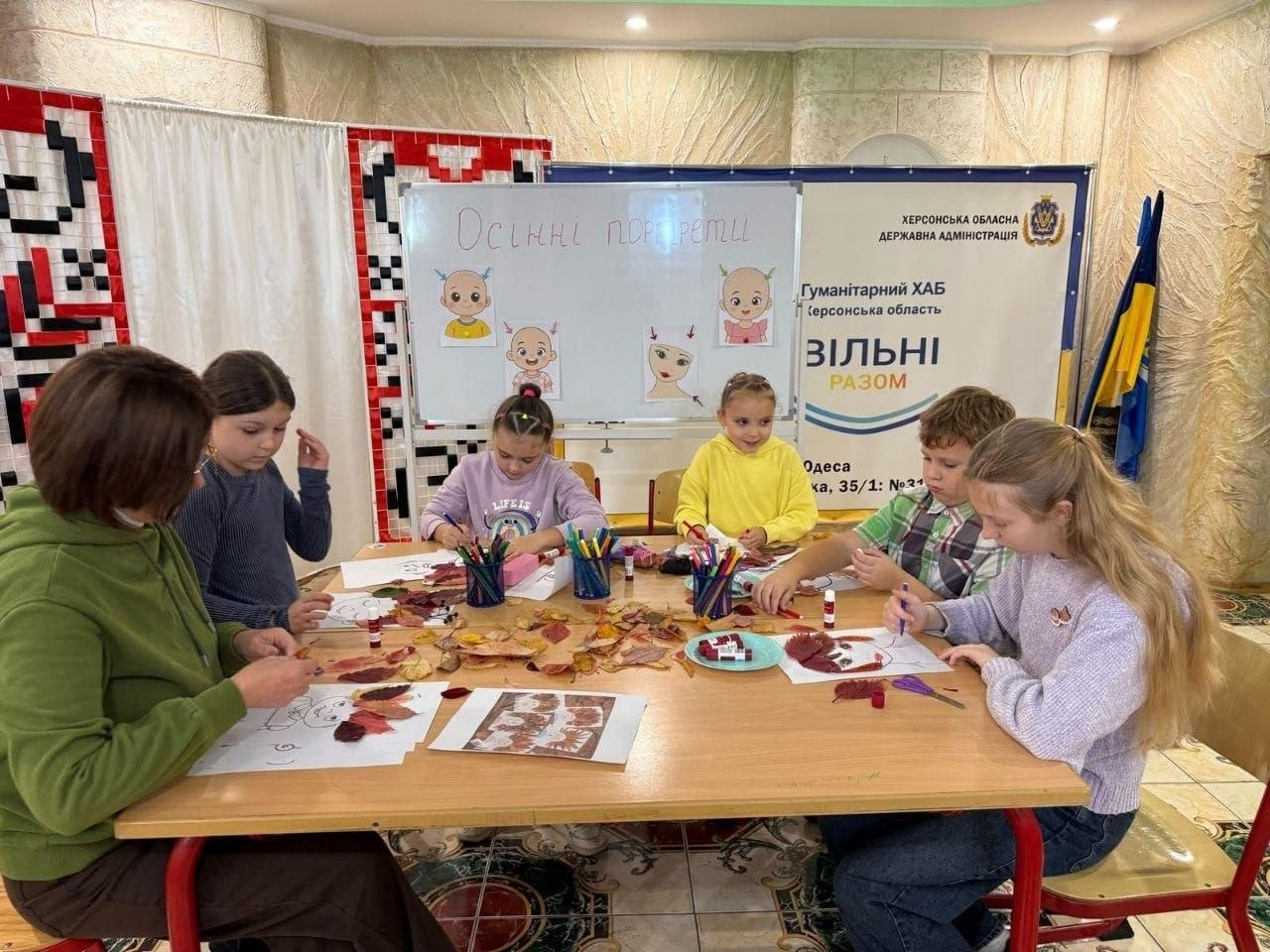 На світлині зображено дітей переселенців із Херсонщини разом із викладачкою під час дитячого майстер-класу «Осінні портрети» в Одеському координаційному центрі «Вільні разом». Вони сидять навколо великого дерев'яного столу, у центрі якого розсипане сухе осіннє листя різних відтінків та стоять стаканчики з кольоровими олівцями й маркерами. З лівого боку столу сидить викладачка в зеленій кофті з капюшоном, а поруч із нею та навколо столу розмістилися хлопчики й дівчатка, які за допомогою клею та заготовок створюють аплікації, де сухе листя виконує роль волосся на намальованих обличчях. За їхніми спинами встановлено білу магнітну дошку з написом крейдою «Осінні портрети» та чотирма зразками дитячих робіт, а ліворуч від неї висить тканинна ширма з традиційним українським червоно-чорним орнаментом. З правого боку кадру позаду столу розміщено великий інформаційний банер із синім фоном, офіційним гербом та текстовим написом «Гуманітарний ХАБ Херсонська область ВІЛЬНІ РАЗОМ Одеса».