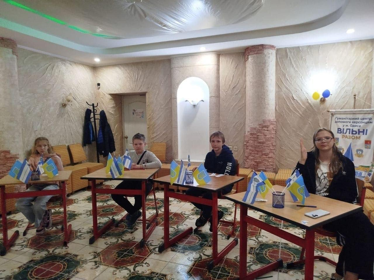 На світлині зафіксовано хід додаткового заняття з української мови для учнів 5–6 класів із родин переселенців із Херсонщини в Одеському координаційному центрі «Вільні разом». Четверо школярів — дві дівчинки та два хлопчики — симетрично розсаджені по одному за окремими одномісними партами, які вишикувані в один ряд серед просторої кімнати. На кожному навчальному столі з червоним металевим каркасом встановлено по два маленькі прапорці: Державний Прапор України та синьо-жовто-синій прапор Херсонської області. Дівчина за крайньою праворуч партою активно підняла руку догори для відповіді, а на її столі поруч із канцелярією лежить смартфон. Простір кабінету з візерунчастою кахельною підлогою та рельєфними колонами доповнює невеликий інформаційний банер «Гуманітарний центр допомоги херсонцям у м. Одеса ВІЛЬНІ РАЗОМ» на задньому плані.