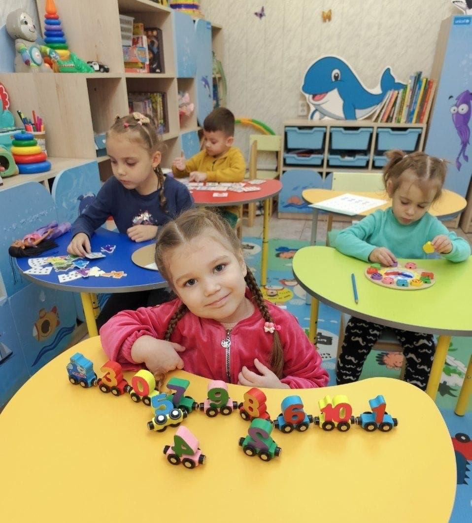 На світлині зображено дітей дошкільного віку з родин внутрішньо переміщених осіб з Херсонщини під час розвивального заняття в Одеському координаційному центрі «Вільні разом». На передньому плані по центру за жовтим фігурним столом сидить маленька усміхнена дівчинка з двома косичками у яскраво-рожевій кофті, яка викладає перед собою іграшковий дерев'яний потяг із різнокольоровими вагонами-цифрами. Позаду неї за сусідніми круглими столами симетрично розсаджені інші діти: ліворуч дівчинка в синій кофті збирає пазли, праворуч дівчинка в бірюзовому светрі розкладає фігурки на спеціальній дошці, а на дальньому плані хлопчик у гірчичній кофті працює з навчальними картками. Заняття проходить у сучасній яскравій ігровій кімнаті з дитячими меблями, малюнком синього дельфіна на шафі для книг, розвивальними пірамідками на полицях та м'яким пазл-килимком із морськими мотивами на підлозі.