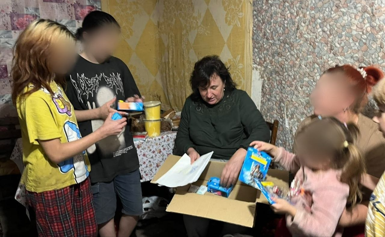 На світлині зафіксовано зустріч начальниці Мирненської селищної військової адміністрації Валентини Головатої з багатодітною родиною, яку повернули з тимчасово окупованих територій. У центрі кадру сидить очільниця МВА Валентина Головата в темно-зеленій кофті з довгими рукавами, яка розглядає дитячі листи біля великої картонної коробки з подарунками та іграшками. З лівого боку від неї стоять двоє підлітків: дівчина в жовтій футболці та кардигані у червоно-чорну клітинку і хлопець у чорній футболці з принтом, які тримають у руках маленькі коробки з солодощами. З правого боку кадру на стільці сидить мати з рудим волоссям, яка тримає на руках маленьку доньку у рожевій пухнастій кофті, що захоплено розглядає нову іграшку в синьому блістерному пакуванні. На самому правому краю кадру видно повернуту в профіль четверту дитину у світлій кофті з дрібним принтом. Обличчя матері та трьох дітей у лівій і центральній частинах кадру штучно розмиті з міркувань безпеки.