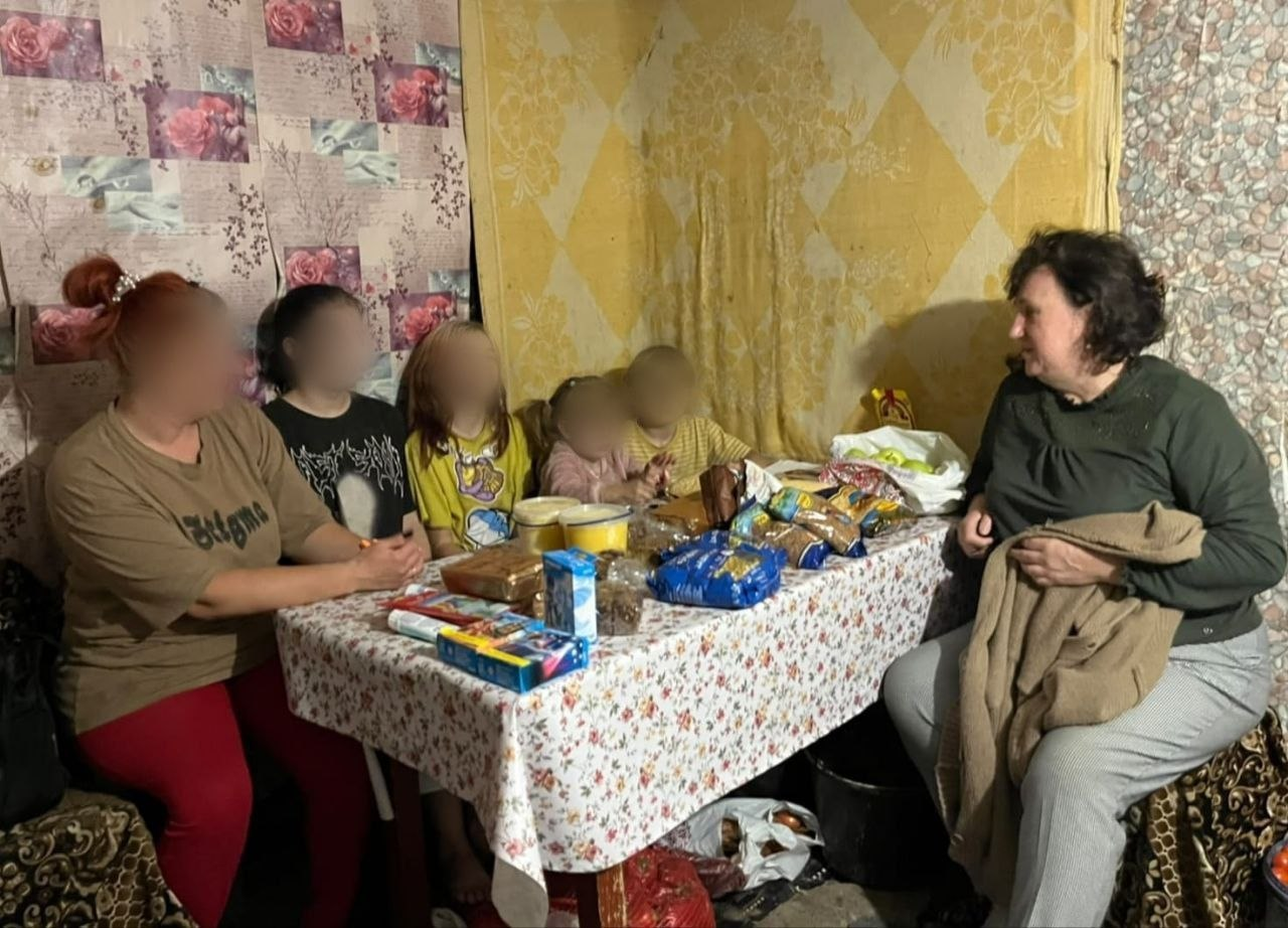 На світлині зафіксовано продовження зустрічі начальниці Мирненської селищної військової адміністрації Валентини Головатої з багатодітною родиною, яку повернули з тимчасово окупованих територій. Праворуч на дивані сидить очільниця МВА Валентина Головата в темно-зеленій кофті та світлих штанях, яка тримає на колінах коричневу куртку й розмовляє з присутніми. Навпроти неї, з лівого боку та по центру за великим столом, розсаджені члени родини: мати у коричневій футболці, хлопець у чорній футболці з принтом, дівчинка в жовтій футболці та двоє найменших дітей у рожевій і жовтій кофтах. Обличчя матері та всіх чотирьох дітей на зображенні штучно розмиті з міркувань безпеки. Весь стіл зі світлою скатертиною в квіточку заставлений гуманітарною допомогою: великими пластиковими відрами з медом, численними прозорими пакунками з крупами та солодощами, коробками з цукерками, печивом та білим пакетом із яблуками.