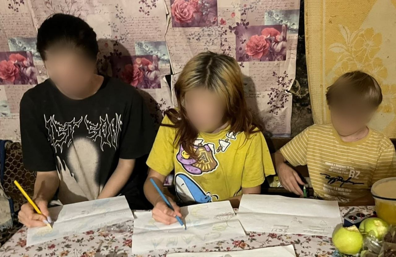 На світлині зафіксовано трьох дітей із багатодітної родини переселенців з Херсонщини, які сидять поруч за столом і зосереджено малюють та пишуть листи до Святого Миколая. Якщо дивитися від правого краю до лівого, першим сидить хлопчик у жовтій смугастій футболці, поруч із ним сидить дівчина в яскравій жовтій футболці з малюнками, яка тримає синій олівець, а ліворуч розмістився старший хлопець у чорній футболці з білим графічним принтом, який пише жовтим олівцем. Перед кожною дитиною на столі зі світлою скатертиною в квіточку розкладено довгі білі паперові аркуші із заготовками малюнків. Обличчя всіх трьох дітей на зображенні штучно розмиті з міркувань безпеки.