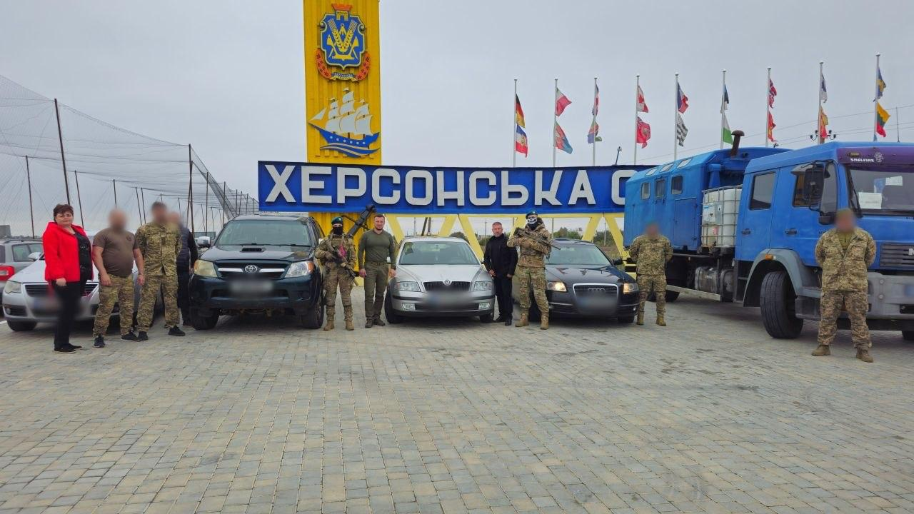 На світлині зображено начальника Херсонської ОВА Олександра Прокудіна, начальницю Милівської СВА Любов Мінько, чоловіка в цивільному одязі та українських військових на тлі стели «ХЕРСОНСЬКА ОБЛАСТЬ» під час передачі автомобілів від литовських партнерів. Учасники заходу стоять на брущатці перед лінійкою наданої техніки: сріблястим седаном Skoda, чорним пікапом Toyota, чорним седаном Audi та великою синьою вантажівкою-автомайстернею. Олександр Прокудін стоїть по центру в мілітарі-одязі безпосередньо біля сріблястого седана Skoda. Любов Мінько в яскравій червоній кофті та чорних штанях стоїть на передньому плані в лівій частині кадру, а далі в ряду розташовані військовослужбовці Сил оборони у камуфляжній формі та чоловік у цивільному одязі. З міркувань безпеки штучно розмито обличчя усіх трьох чоловіків біля Любові Мінько, а також обличчя двох військових із правого боку кадру. Над синьо-жовтим написом стели височіє герб Херсонщини з вітрильником, а праворуч на флагштоках майорять прапори України та Литви.