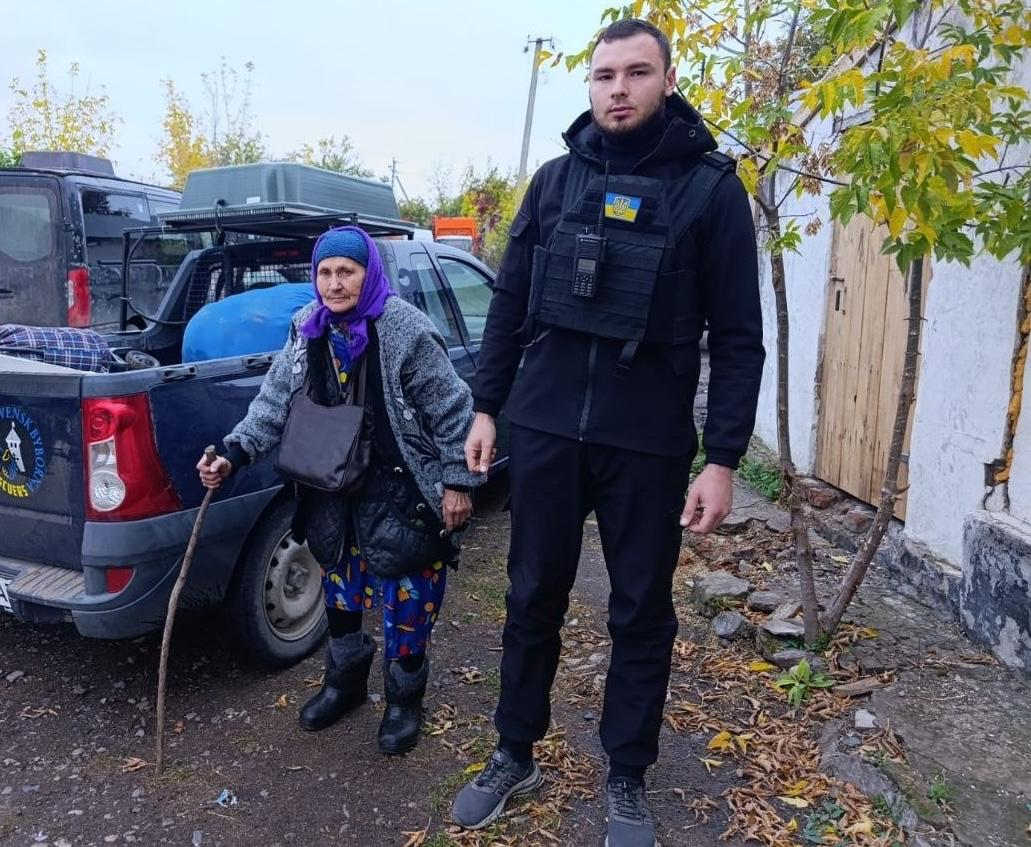 На світлині зображено волонтера Андрія Романіва та 85-річну жительку під час її евакуації з прифронтового села Урожайне Бериславської громади. Праворуч стоїть молодий волонтер у чорному тактичному одязі та бронежилеті з українським прапором і рацією. Ліворуч біля нього стоїть літня жінка у фіолетовій хустці, теплій куртці та сірій кофті, яка спирається на дерев'яну палицю. Вони перебувають на подвір'ї біля синього пікапа, у відкритому кузові якого складено великі пакунки з речами евакуйованої жінки. На задньому плані видно білу стіну сільського будинку та осінні дерева з жовтим листям.