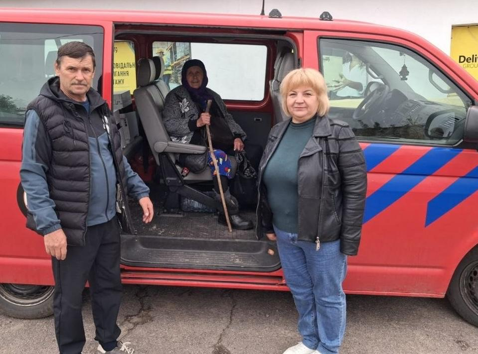 На світлині зображено наступний етап евакуації 85-річної жительки села Урожайне на тлі червоного мікроавтобуса з синіми смугами. Літня жінка у фіолетовій хустці вже сидить у салоні автомобіля біля відкритих розсувних дверей, тримаючи в руках свою дерев'яну палицю. Перед машиною стоять двоє людей: ліворуч — чоловік у чорній безрукавці поверх сірой кофти та темних штанях, а праворуч — жінка зі світлим волоссям у чорній шкіряній куртці, темно-зеленому гольфі та блакитних джинсах. На задньому плані помітна частина білої будівлі з елементом жовтої вивіски.