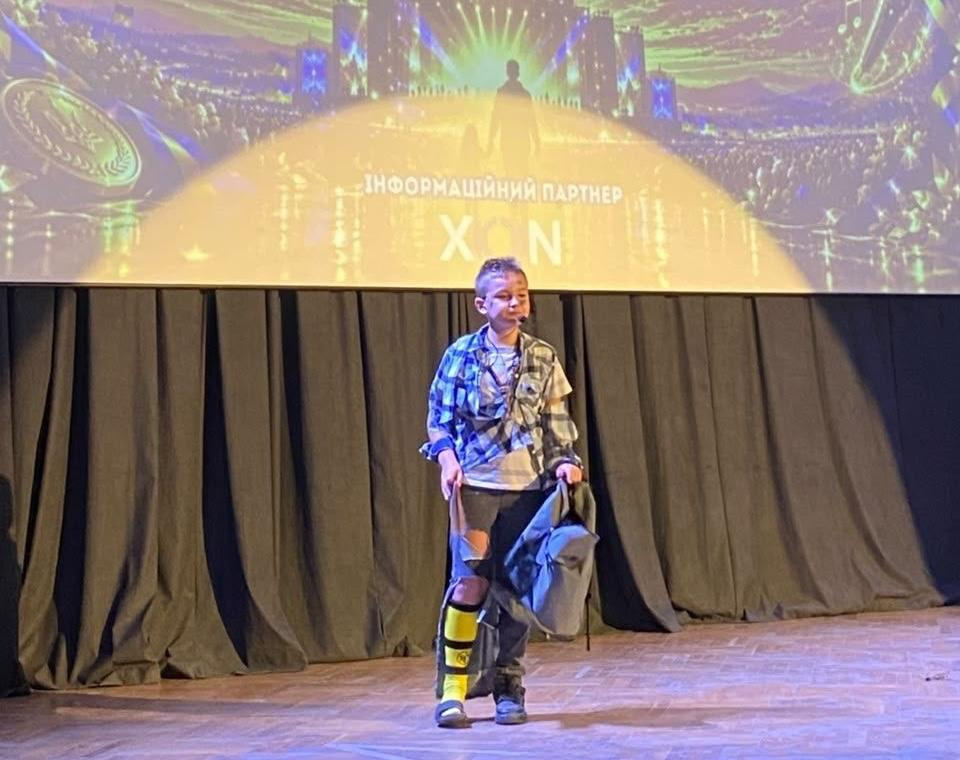 На світлині зафіксовано сольний виступ хлопчика на сцені під час фіналу творчого фестивалю «Мистецький шлях» у Львівському палаці мистецтв. Юний актор стоїть по центру дерев'яного помосту в сценічному образі: на ньому вдягнена розстебнута синьо-сіра картата сорочка поверх білої футболки, порвані темні джинси, а на правій нозі виділяється яскраво-жовтий гольф із чорними смугами. У лівій руці він тримає сірий рюкзак, а до його обличчя прикріплено маленький наочний мікрофон-гарнітуру. Задній план сцени завішений суцільною темною тканиною, над якою височіє великий мультимедійний екран із золотистою заставкою фестивалю.