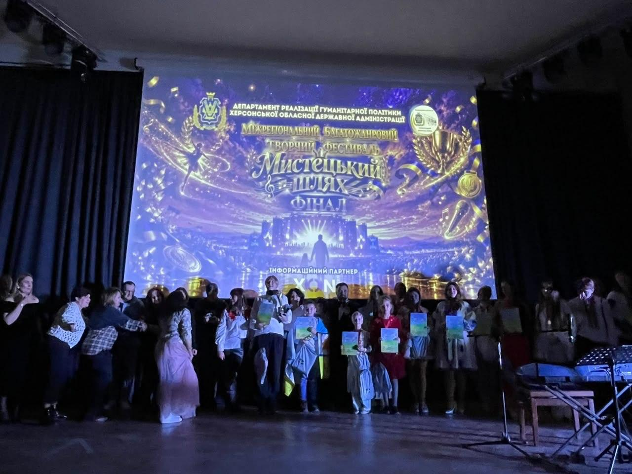 На світлині зафіксовано фінальну частину та нагородження учасників Міжрегіонального багатожанрового творчого фестивалю «Мистецький шлях» на сцені Львівського палацу мистецтв. Посередині кадру безпосередньо за одним із хлопчиків стоїть директорка Департаменту реалізації гуманітарної політики ХОДА Віолета Царькова. Поруч, праворуч від неї, за іншим хлопчиком стоїть заступник директора Департаменту реалізації гуманітарної політики ХОДА Андрій Ярьоменко. Велика група талановитих дітей, підлітків та дорослих виконавців вишикувалася в один ряд на темному дерев'яному помості сцени, тримаючи в руках яскраві блакитно-жовті дипломи та нагороди фіналістів. Присутні одягнені у різноманітні сценічні та патріотичні костюми. Весь задній план займає величезний мультимедійний екран із яскравою золотисто-синьою заставкою, де розміщено герб Херсонщини, зображення кубків, медалей та написи «ДЕПАРТАМЕНТ РЕАЛІЗАЦІЇ ГУМАНІТАРНОЇ ПОЛІТИКИ ХЕРСОНСЬКОЇ ОБЛАСНОЇ ДЕРЖАВНОЇ АДМІНІСТРАЦІЇ», «Мистецький шлях. ФІНАЛ». Ліворуч і праворуч сцену обрамляють чорні куліси, а у правому кутку помітно музичне обладнання та інструменти.