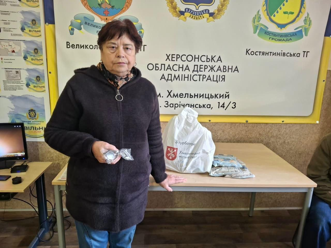 На світлині зафіксовано жінку старшого віку з числа внутрішньо переміщених осіб з Херсонщини, яка отримує допомогу у гуманітарному центрі «Вільні разом» у місті Хмельницький. Вона стоїть на передньому плані ліворуч у темному довгому пальті та синіх джинсах, тримаючи в руках блістерні упаковки з медикаментами. Позаду неї розташовано довгий дерев'яний стіл, на якому лежить великий білий пакет із продуктами та червоним логотипом «Хмельницька міська рада», а поруч розкладено сірі прозорі пакунки з рисовою сумішшю. Робоче місце облаштоване на тлі великого жовтого офіційного стенда з офіційними написами «ХЕРСОНСЬКА ОБЛАСНА ДЕРЖАВНА АДМІНІСТРАЦІЯ м. Хмельницький, а також гербами Великокопанівської ТГ та Костянтинівської ТГ. Ліворуч у кадрі помітно частину комп'ютерного монітора на робочому столі волонтерів, а праворуч на задньому плані сидить інший відвідувач центру в зеленій кофті.