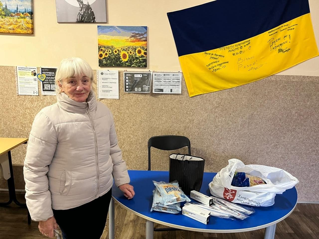 На світлині зафіксовано жінку старшого віку з числа внутрішньо переміщених осіб з Херсонщини, яка отримує допомогу у Хмельницькому гуманітарному центрі «Вільні разом». Вона стоїть у лівій частині кадру в світло-бежевій куртці біля круглого синього столу, на якому розкладено гуманітарні пакунки. На столі лежать кілька упаковок рисової суміші, білі коробки з медикаментами, чорний паперовий пакет та великий білий пакет із продуктами, на якому нанесено червоний логотип «Хмельницька міська рада». На задньому плані на стіні розвішані дитячі малюнки із соняшниками, інформаційні плакати та оголошення центру, а праворуч закріплено великий синьо-жовтий Державний Прапор України з патріотичними підписами та написами «Разом до Перемоги!» та «Вільні Разом!».