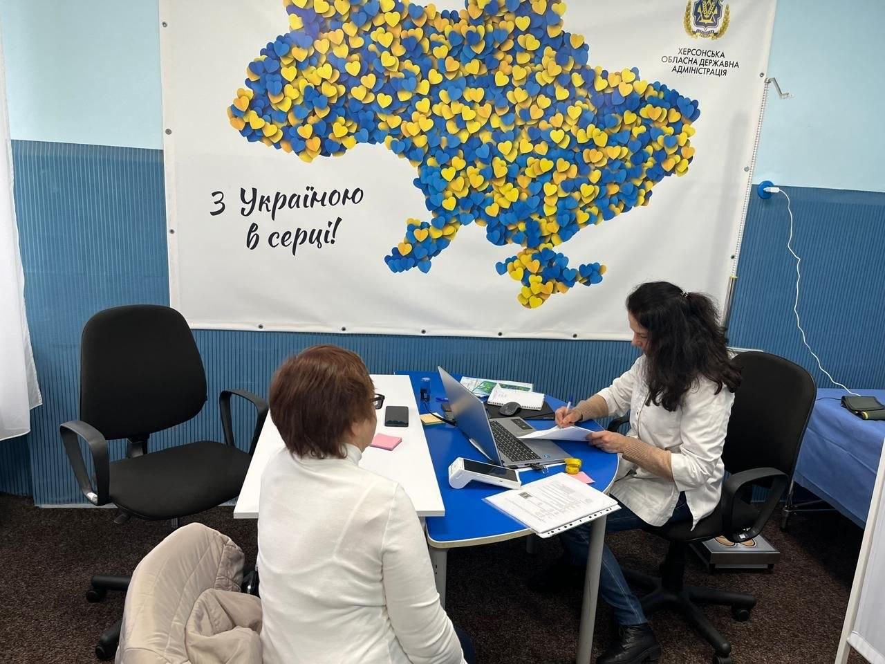 На світлині зафіксовано робочий момент надання медичної допомоги у Хмельницькому гуманітарному центрі «Вільні разом». Праворуч за круглим синім столом сидітиме представниця мобільної бригади Товариства Червоного Хреста України в білій сорочці та синіх джинсах, яка робить записи у документах та працює за відкритим ноутбуком. Навпроти неї на стільці спиною до об'єктиву сидить жінка-переселенка у світлій кофті, яка прийшла на медичну консультацію. На столі перед фахівчинею розкладено папери, бланки, лежить платіжний термінал та канцелярія. Робоче місце облаштоване у кабінеті з блакитними стінами на тлі великого білого банера із зображенням мапи України, що викладена з маленьких синьо-жовтих сердечок, написом «З Україною в серці!» та офіційним логотипом «ХЕРСОНСЬКА ОБЛАСНА ДЕРЖАВНА АДМІНІСТРАЦІЯ» у верхньому кутку.