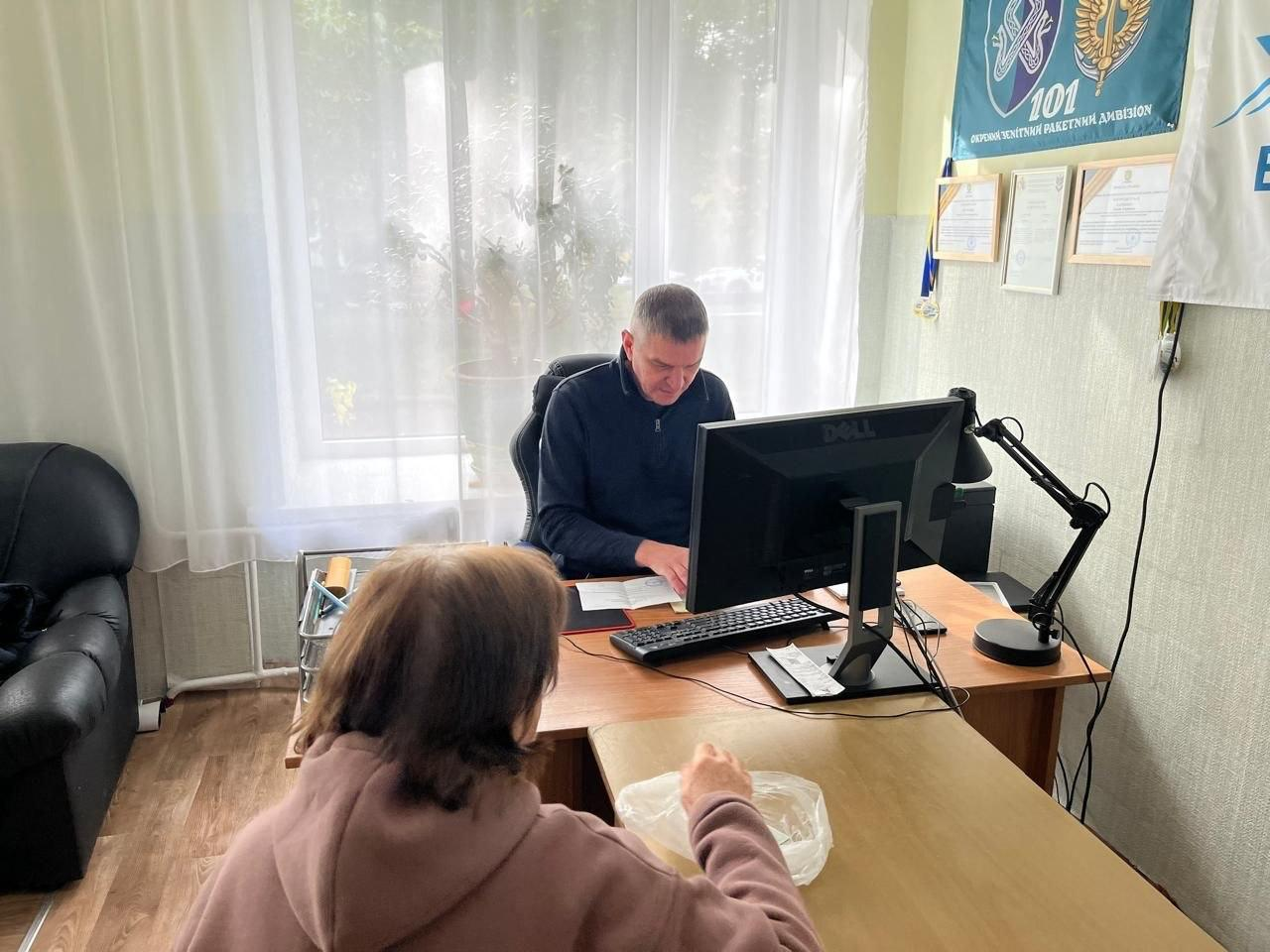 На світлині зафіксовано робочий момент надання консультаційних послуг у Хмельницькому гуманітарному центрі «Вільні разом». За великим дерев'яним столом у центрі кадру сидить чоловік-консультант у темно-синій кофті, який вивчає відкритий документ відвідувачки. Перед ним на столі встановлено монітор комп'ютера, стоїть настільна лампа та лежить клавіатура. На передньому плані спиною до об'єктиву сидить жінка у коричневому худі, яка звернулася по допомогу. Кабінет облаштований світлими стінами та лінолеумом під дерево, ліворуч стоїть чорний шкіряний диван, а позаду консультанта розташоване велике вікно з білою прозорою тюлею. На стіні праворуч розміщено інформаційні сертифікати у рамках, а вгорі закріплено бірюзовий прапор із символікою та написом «101 ОКРЕМІЙ ЗЕНІТНИЙ РАКЕТНИЙ ДИВІЗІОН».
