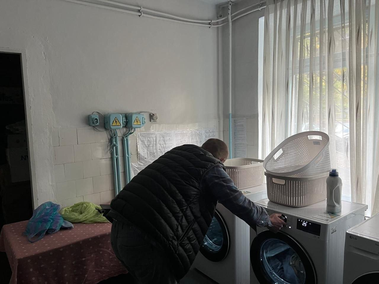 На світлині зафіксовано процес роботи соціальної пральні у Хмельницькому гуманітарному центрі «Вільні разом». У центрі кадру спиною до об'єктиву стоїть чоловік у темній жилетці поверх сорочки, який схилився над білою пральною машиною та налаштовує круглий перемикач програм на цифровій панелі керування. Поруч у ряд встановлено ще дві білі пральні та сушильні машини. На кришці однієї з них стоять три великі пластикові кошики для білизни білого та рожевого кольорів, а також пляшка рідкого засобу для прання. Ліворуч у кадрі помітно стіл із рожевою скатертиною, на якому складено кольорові текстильні речі. Приміщення пральні має білі стіни з ділянками світлої кахельної плитки, захисні блоки електромережі з позначками вольтів, а велике вікно праворуч завішене світлою напівпрозорою тюлею.