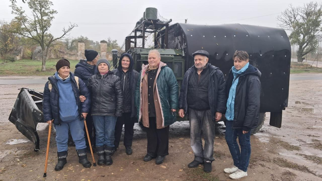 На світлині зафіксовано групу евакуйованих жителів Берислава, серед яких п'ять жінок та два літні чоловіки, які стоять разом із волонтеркою на тлі великого військового евакуаційного автомобіля. Учасники заходу вишикувалися в один ряд на розмоклій від дощу ґрунтовій дорозі та одягнені у теплий осінній одяг: куртки, пальта, шапки, кепки та чоботи, а крайня ліворуч жінка спирається на дві дерев'яні палиці. Позаду них стоїть спеціалізована військова вантажівка темно-зеленого кольору, задня частина якої обладнана масивним напівкруглим чорним захисним тентом-капсулою. Операція проходила у похмурий день на відкритій місцевості прифронтової зони, де на задньому плані помітно асфальтовану дорогу, залишки цегляного паркану та поодинокі дерева без листя.