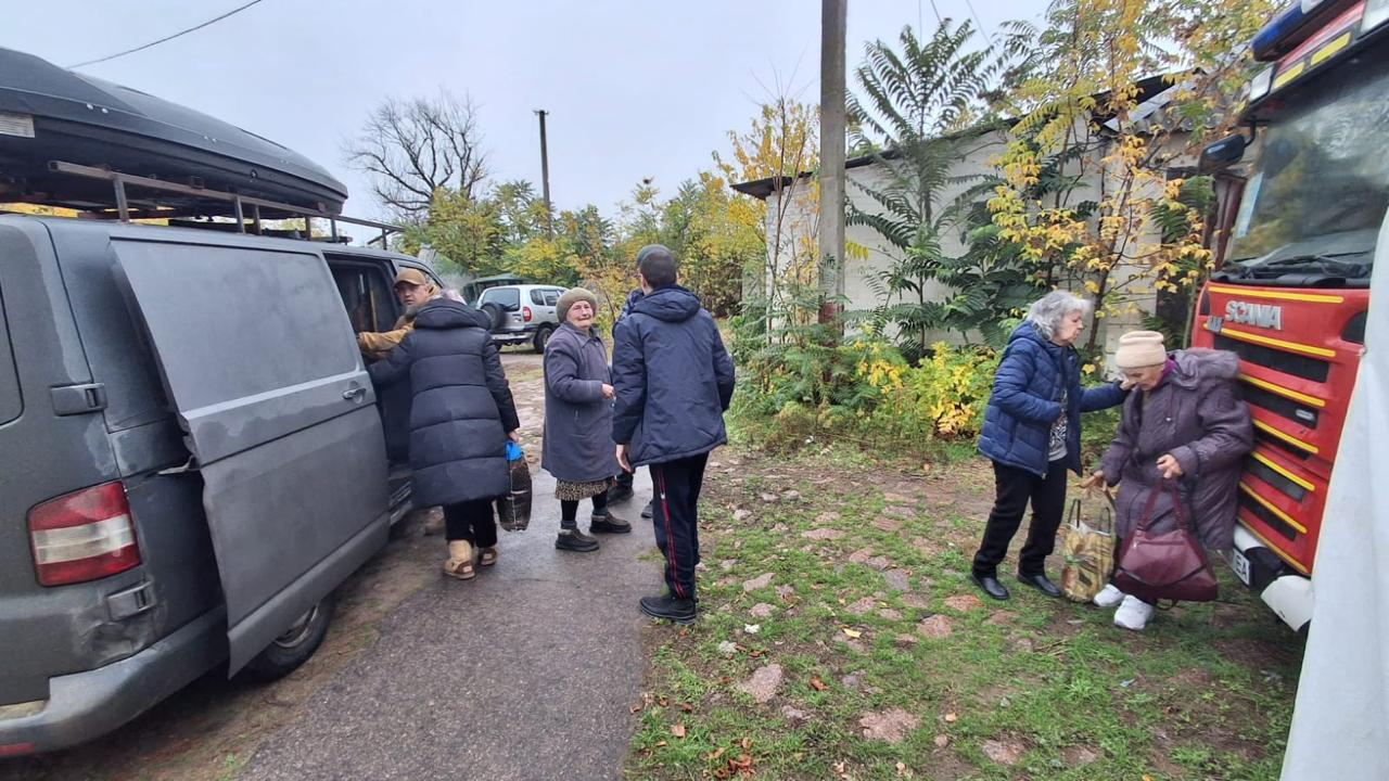 На світлині зафіксовано процес посадки евакуйованих мешканців Берислава у транспорт на проміжному пункті збору. Ліворуч на передньому плані стоїть сірий мікроавтобус Volkswagen із відкритими боковими зсувними дверима та встановленим на даху додатковим пластиковим багажником-боксом. Біля машини спиною до об'єктиву стоїть літня жінка у довгому темному пуховику, якій чоловік усередині салону допомагає сісти на пасажирське місце. Поруч по центру кадру розмовляють літня жінка у сірому пальті й хустці та молодий хлопець у синій куртці, який стоїть спиною, а безпосередньо за його спиною майже непомітно волонтера Андрія Петухова на позивний «Боксер» у касці. Праворуч видно передню частину великої червоної рятувальної вантажівки, біля радіаторної решітки якої стоять дві літні жінки з пакунками та сумками, підтримуючи одна одну. Подія розгортається на подвір'ї з ґрунтово-трав'яним покриттям на тлі одноповерхових прибудов та зелених кущів під похмурим небом.