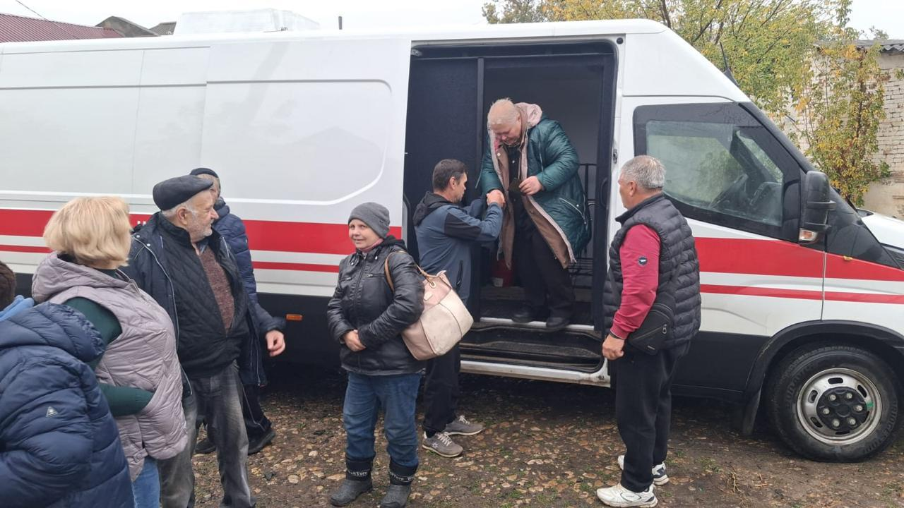 На світлині зафіксовано процес висадки евакуйованих жителів Берислава біля великого білого мікроавтобуса з червоною горизонтальною смугою на кузові. У центрі кадру з відкритих бокових зсувних дверей автомобіля обережно виходить літня жінка у блискучій зеленій куртці, якій допомагає спуститися чоловік у синій кофті, підтримуючи її за руку. Навколо них на землі стоять інші врятовані мешканці та волонтери. Праворуч на передньому плані боком до об'єктиву стоїть сивочолий чоловік у червоній кофті та чорній дутій жилетці. Ліворуч розмістилася група людей, серед яких виділяється літній чоловік у чорній кепці та темній куртці, а поруч із ним стоїть жінка у сірій шапці та чорній шкіряній куртці з бежевою сумкою через плече. Подія розгортається на ґрунтовому подвір'ї у похмурий день, а на задньому плані видно частину даху сусідньої будівлі та осінні дерева з жовтіючим листям.