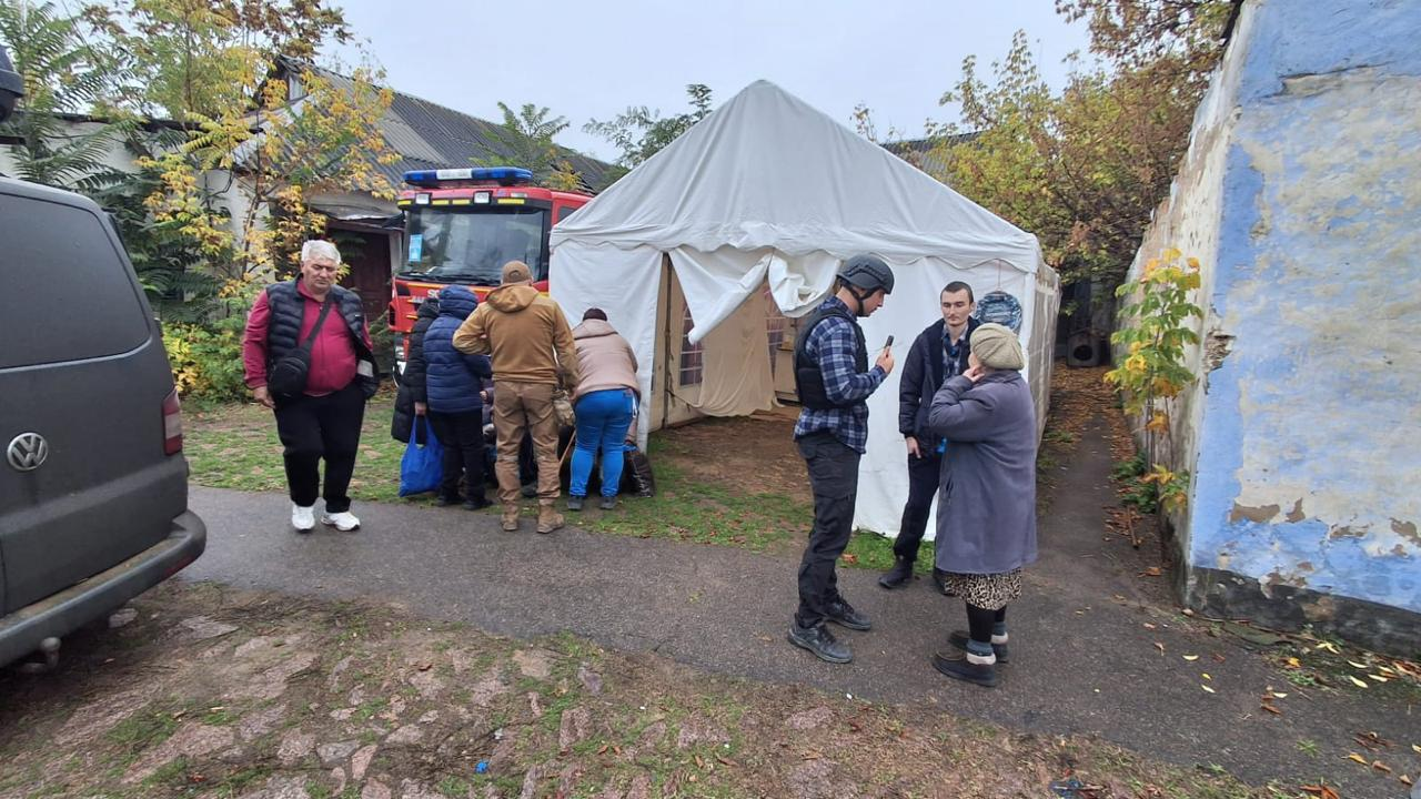 На світлині зафіксовано транзитний пункт збору або проміжну зупинку під час евакуаційної операції жителів Берислава. У центрі кадру встановлено великий білий армійський намет, біля входу до якого праворуч стоять і спілкуються троє людей: волонтер Андрій Петухов на позивний «Боксер» у касці, бронежилеті та картатій сорочці, молодий хлопець у куртці та літня жінка у сірому пальті й хустці. Ліворуч від намету інша група евакуйованих жителів розбирає свої речі та сині сумки біля великої червоної рятувальної машини ДСНС. На лівому краю кадру на передньому плані видно частину задньої сторони сірого мікроавтобуса Volkswagen, біля якого стоїть сивочолий чоловік у червоній жилетці поверх бордової кофти та чорних спортивних штанях. Дія відбувається на подвір'ї зі старою брущаткою, де праворуч височіє облуплена синьо-біла стіна будівлі, а на задньому плані ростуть осінні дерева з пишною зелено-жовтою кроною.
