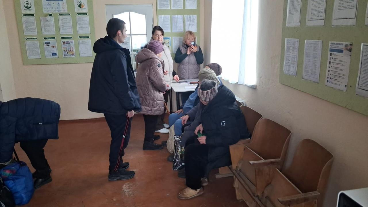 На світлині зафіксовано групу евакуйованих жителів Берислава всередині приміщення транзитного пункту або центру оформлення документів. У лівій частині кадру стоїть молодий хлопець у чорній куртці та спортивних штанях, який спілкується з літньою жінкою в рожевій зимовій куртці та хутці. Поруч із ними за маленьким столом перебувають дві соціальні працівниці у світлих жилетках, одна з яких фіксує процес на смартфон. Праворуч на дерев'яних відкидних стільцях та лавці вздовж світлої стіни відпочивають інші евакуйовані літні жінки в теплих хустках і куртках. Приміщення має коричневу підлогу, велике вікно з білою тюлею на задньому плані, а стіни облаштовані великими зеленими інформаційними стендами, на яких розвішані численні бланки, пам'ятки та паперові документи.