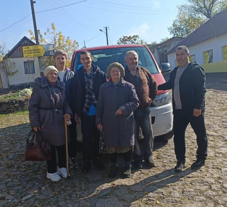 На світлині у повний зріст зображено групу евакуйованих жителів Берислава разом із супроводжуючими особами під час спільного фото на тлі червоного мікроавтобуса. Учасники заходу вишикувалися в один ряд на брущатому подвір'ї у сонячний день: ліворуч стоїть літня жінка у сірому довгому пальті й хустці, яка спирається на дерев'яну палицю, за нею стоїть жінка зі світлим волоссям, по центру розташовані молодий хлопець у темній куртці й картатій сорочці та ще одна літня жінка у сірому пальті й пишній шапці. Праворуч від них стоїть літній чоловік у коричневому в'язаному светрі та сірих джинсах, а на самому правому краю кадру чоловік у чорній спортивній куртці та штанях тримається рукою за капот автомобіля. На задньому плані видно одноповерхові сільські будинки, лінію електропередач та осінні дерева з жовто-зеленим листям.