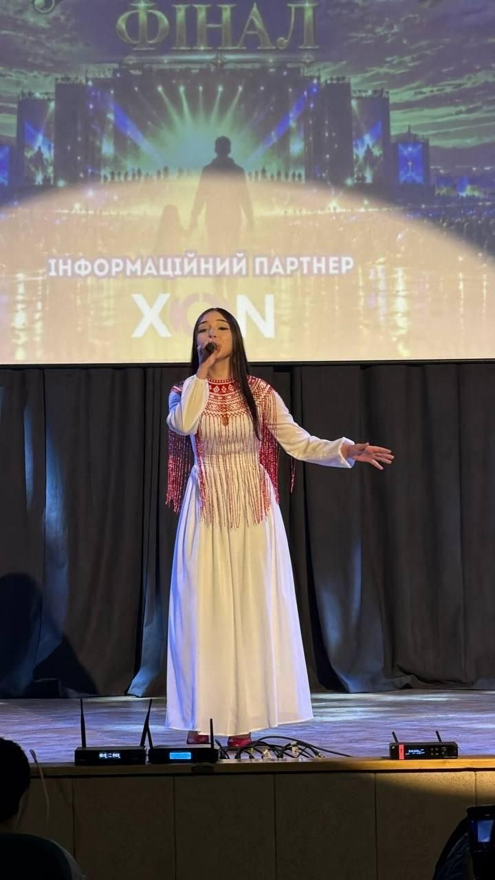 На світлині зафіксовано сольний виступ вокалістки Марії Богдан на сцені Львівського палацу мистецтв під час виконання нової пісні про Херсонщину на гала-концерті фестивалю «Мистецький шлях». Співачка з довгим темним волоссям стоїть по центру дерев'яного помосту в довгій білій сукні, верхня частина якої прикрашена масивним патріотичним орнаментом та довгими червоними кораловими нитками-торочками на плечах і грудях. Вона тримає правою рукою мікрофон біля обличчя, а ліву руку емоційно відвела вбік. Задній план сцени завішений суцільною темною тканиною, над якою встановлено великий мультимедійний екран із золотистою заставкою заходу, де вгорі помітний напис «ФІНАЛ», а по центру — силует людини на сцені та текст «ІНФОРМАЦІЙНИЙ ПАРТНЕР XON». На передньому плані внизу кадру видно комутаційне та звукове обладнання на краю сцени.