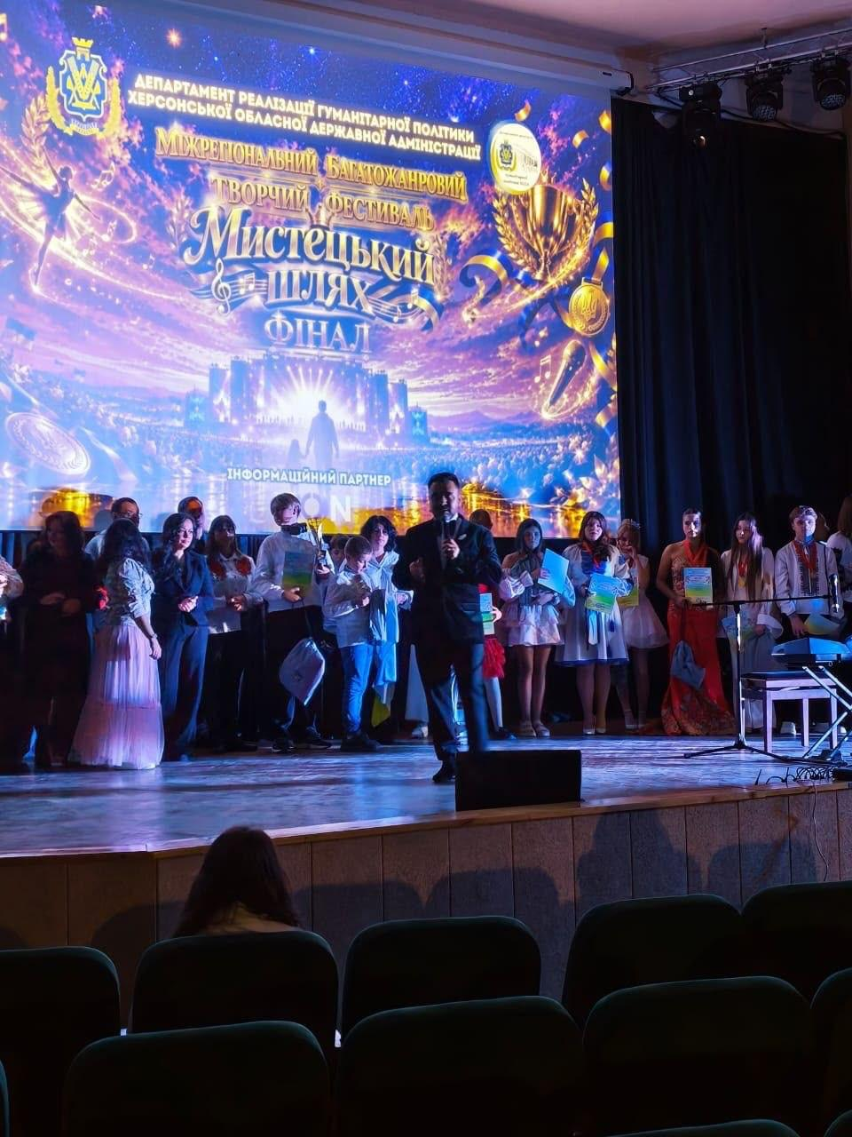 На світлині зафіксовано виступ ведучого на сцені під час фіналу та нагородження учасників Міжрегіонального багатожанрового творчого фестивалю «Мистецький шлях». По центру сцени на передньому плані стоїть заступник директора Департаменту реалізації гуманітарної політики ХОДА Андрій Ярьоменко у класичному чорному костюмі з мікрофоном у руці, який виступає в ролі ведучого та звертається до глядацької зали. За його спиною в один ряд вишикувалися численні учасники фестивалю — діти та підлітки у сценічних костюмах і вишиванках, які тримають у руках свої дипломи та нагороди. Весь задній план сцени займає величезний мультимедійний екран із яскравою золотистою заставкою, де розміщено герб Херсонщини та написи «ДЕПАРТАМЕНТ РЕАЛІЗАЦІЇ ГУМАНІТАРНОЇ ПОЛІТИКИ ХЕРСОНСЬКОЇ ОБЛАСНОЇ ДЕРЖАВНОЇ АДМІНІСТРАЦІЇ», «Мистецький шлях. ФІНАЛ» та «ІНФОРМАЦІЙНИЙ ПАРТНЕР XON». Перед сценою внизу кадру видно темні ряди крісел та голови глядачів у залі.