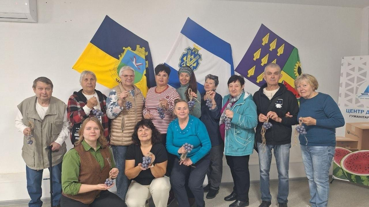 На світлині зафіксовано групове фото учасників творчого майстер-класу в Черкаському гуманітарному центрі «Вільні разом», які тримають у руках виготовлені власноруч грона винограду з фіолетової пряжі. Відвідувачі старшого віку та майстрині-волонтери позують у залі на тлі білої стіни, де в один ряд закріплені три прапори: ліворуч — Державний Прапор України з гербом Херсонщини, посередині — прапор Херсонської області, а праворуч — прапор із жовтими наконечниками стріл та соняшником. Частина жінок на передньому плані присіла навпочіпки та нахилилася, а решта учасників вишикувалися в один ряд позаду них. На правому краю кадру помітно частину брендованого стенда гуманітарного хабу із зображенням кавуна.