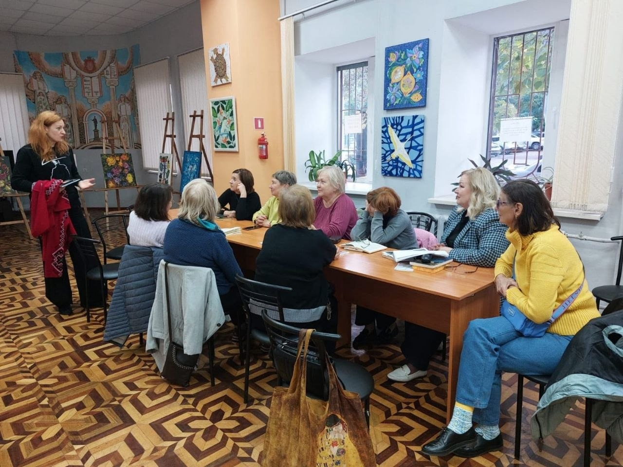 На світлині зафіксовано літературну зустріч для внутрішньо переміщених осіб з Херсонщини в Одесі, яку організувала публічна бібліотека Верхньорогачицької селищної ради. У залі за довгим дерев'яним столом півколом сидить група жінок-переселенок старшого віку, які уважно слухають доповідачку. Ліворуч біля столу стоїть пані Неля у чорному одязі з червоною хусткою на руці, яка виступає перед присутніми. Учасниці зустрічі одягнені у повсякденний одяг: кофти, светри та піджаки різних кольорів, зокрема жінка на передньому плані праворуч сидить у яскраво-жовтій кофті та синіх джинсах. Захід проходить у залі з фігурним паркетом, світло-блакитними стінами та великими вікнами, між якими розвішані картини, а на задньому плані ліворуч на мольбертах виставлено інші художні роботи.