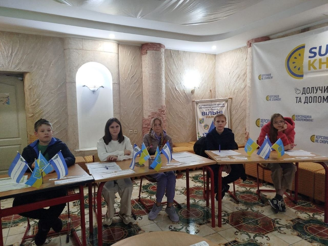 На світлині зображено юних переселенців із Херсонщини під час заняття з української мови у гуманітарному центрі «Вільні разом» у місті Одеса. П'ятеро дітей (два хлопчики та три дівчинки) розсаджені півколом за навчальними столами у світлій кімнаті з декоративними колонами та візерунчастою плиткою на підлозі. Перед ними на столах розкладено папери із завданнями, ручки та виставлено маленькі прапорці України та Херсонської області. На задньому плані між колонами встановлено невеликий брендований банер із гербом та написом «Гуманітарний центр допомоги херсонцям у м. Одеса ВІЛЬНІ РАЗОМ», а праворуч височіє великий інформаційний стенд із написами «ДОЛУЧАЙСЯ ТА ДОПОМАГАЙ», «ХЕРСОНЩИНА ЦЕ НЕЗЛАМНІ!» та іншими патріотичними гаслами. Дві дівчинки та хлопчик ліворуч уважно дивляться в об'єктив, тоді як хлопчик праворуч тримає ручку біля паперів, а крайня праворуч дівчинка у рожевій кофті спирається підборіддям на руку.