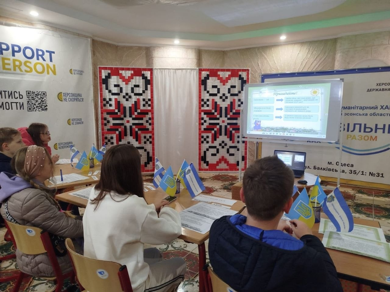На світлині зображено інший ракурс уроку для юних переселенців із Херсонщини в одеському гуманітарному хабі «Вільні разом». Діти сидять за навчальними столами обличчям до великого телевізійного екрана, на якому відображається інтерактивний слайд презентації про роди іменників із заголовком «Пригадайте!». Попереду, спиною до об'єктиву, сидять хлопчик у синій кофті та дівчинка у білому світшоті, а ліворуч за сусіднім столом видно інших дітей, зокрема дівчинку в окулярах та дівчинку в рожевому обручі. На столах перед кожним розкладено навчальні листи та виставлено прапорці України і Херсонської області. Задній план зали прикрашений двома вертикальними полотнами з великим червоно-чорним українським вишитим орнаментом, ліворуч розташований банер проєкту із QR-кодом, а праворуч на стіні нанесено напис із адресою хабу.
