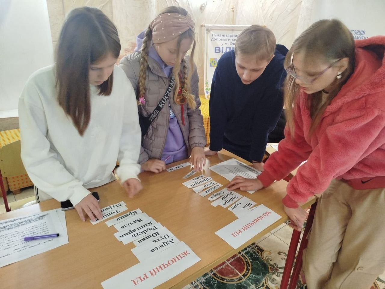 На світлині зображено практичну частину ігрового заняття з української мови для дітей-переселенців у гуманітарному центрі «Вільні разом» в Одесі. Четверо юних учасників (дві дівчинки ліворуч, хлопчик та дівчинка в окулярах праворуч) схилилися над великим дерев'яним столом, де вони розсортовують паперові картки зі словами за трьома категоріями. На столі перед ними лежать великі друковані аркуші-вказівники «ЖІНОЧИЙ РІД» та «ЧОЛОВІЧИЙ РІД», під якими діти викладають слова (зокрема «Адреса», «Путь», «Юшка», «Біль», «Одяг», «Дніпро»). На задньому плані помітно частину світлої стіни кімнати з декоративною колоною та невеликим плакатом хабу.