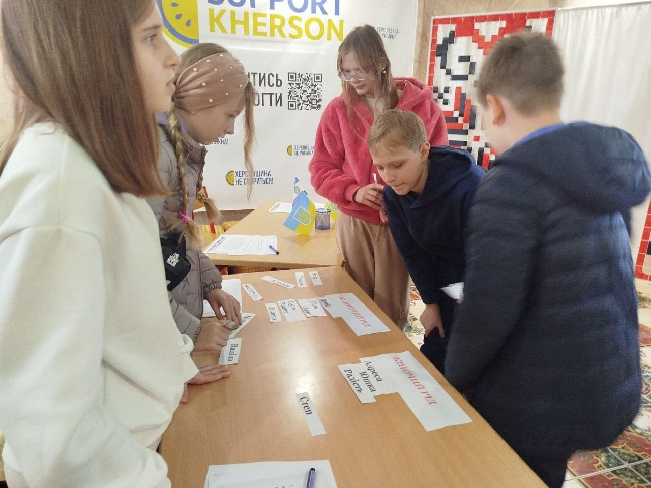На світлині зображено інший момент практичного ігрового завдання з української мови в гуманітарному центрі «Вільні разом» в Одесі. П'ятеро юних переселенців із Херсонщини (три дівчинки ліворуч та два хлопчики праворуч) згрупувалися навколо столу, де вони розкладають паперові картки зі словами за родами іменників. На столі перед ними лежать підписи «ЖІНОЧИЙ РІД» та «ЧОЛОВІЧИЙ РІД», під якими розміщено картки зі словами «Адреса», «Юшка», «Радість», «Степ», «Валіза», «Дніпро», «Біль» та іншими. Задній план прикрашений великим білим банером проєкту SUPPORT KHERSON із QR-кодом та патріотичними наліпками, а праворуч видно частину полотна з традиційним червоно-чорним українським вишитим орнаментом.
