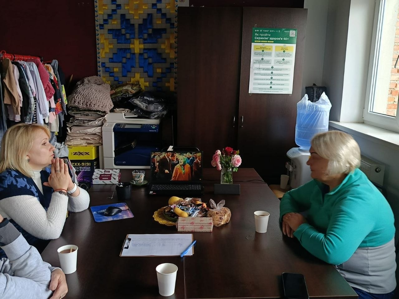 На світлині зображено інший ракурс засідання Чайного розмовного клубу для переселенок із Херсонщини в координаційному центрі «Вільні разом» в Івано-Франківську. Навколо великого дерев'яного столу сидять і спілкуються три жінки, а від четвертої учасниці в лівому нижньому кутку видно лише частину рук і теплу сіру кофту. Ліворуч за столом сидить жінка у синій жилетці з візерунком «гусяча лапка» поверх білого гольфа, яка тримає складені долоні біля обличчя та уважно слухає співрозмовницю. Навпроти неї праворуч сидить сивочола жінка у бірюзово-сірій флісовій кофті, поклавши руки на стіл. По центру столу встановлено відкритий ноутбук, на екрані якого зафіксовано кадр із документального фільму, поруч стоїть маленька скляна ваза з рожевими трояндами, тарілка з частуваннями та паперові стаканчики. На задньому плані помітно велику темну шафу з медичним плакатом «Як пройти скринінг здоров'я 40+», великий синьо-жовтий в'язаний плед або панно, офісний принтер, бутель із водою та вішак із різноманітним одягом у лівому кутку кімнати.