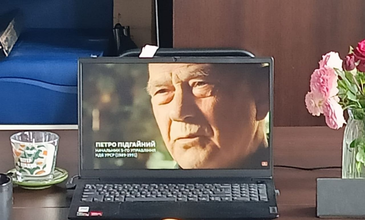 На світлині крупним планом зображено чорний ноутбук, встановлений на коричневому дерев'яному столі під час перегляду документального фільму в гуманітарному центрі «Вільні разом» в Івано-Франківську. На екрані лептопа зафіксовано кадр із літнім чоловіком та чітким текстовим титром зеленого і білого кольорів: «ПЕТРО ПІДГАЙНИЙ НАЧАЛЬНИК 5-ГО УПРАВЛІННЯ КДБ УРСР (1989-1991)». Праворуч від ноутбука на столі стоїть прозора скляна ваза з пишними рожевими трояндами, а ліворуч помітно великий прозорий скляний свічник із зелено-помаранчевим візерунком та край чорного кухля. На задньому плані кадру видніється частина темної офісної шафи та синій елемент меблів або обладнання.