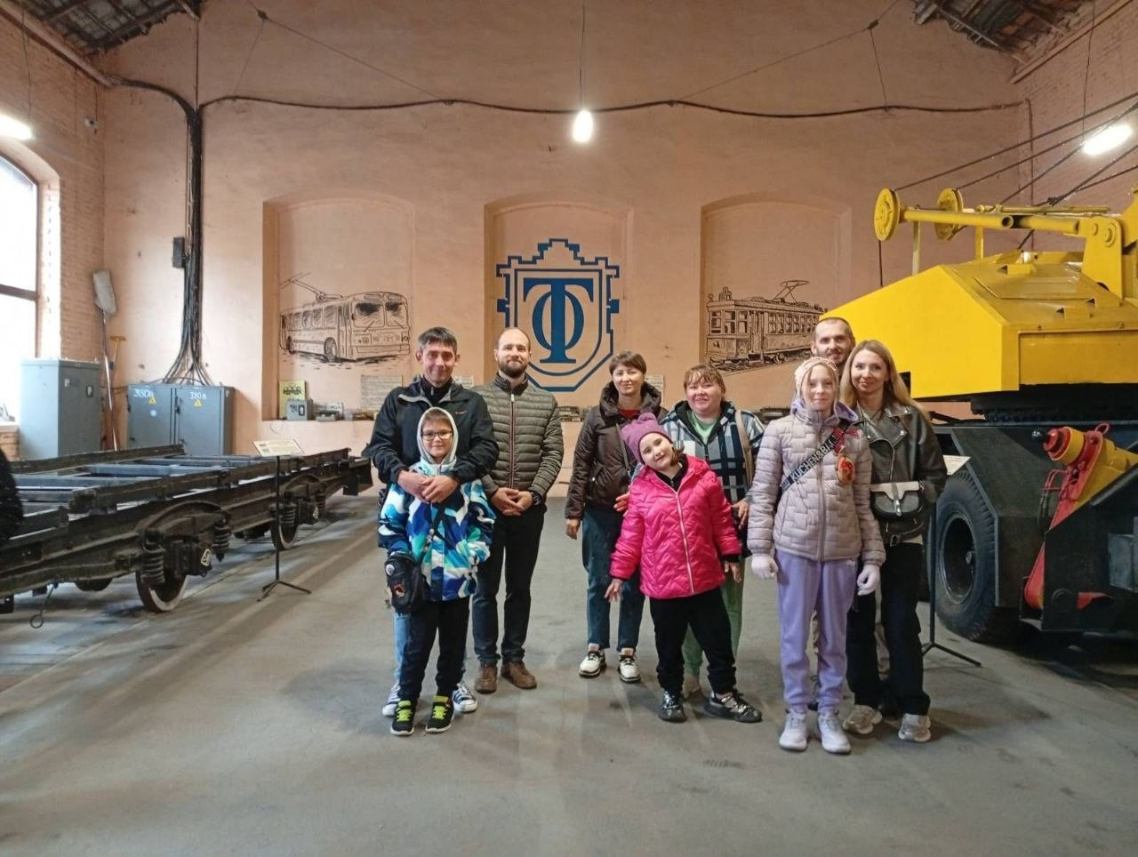 На світлині зображено спільне фото родин внутрішньо переміщених осіб із Херсонщини під час екскурсії до Музею електротранспорту в Одесі. Група з п'яти дорослих та трьох дітей позує в центрі просторого музейного ангара з високою стелею. Присутні одягнені в осінні куртки та куртки-пуховики різних кольорів. На передньому плані ліворуч стоїть чоловік, який тримає за плечі хлопчика в яскравому синьо-білому куртці, а праворуч по центру посміхається дівчинка у рожевій куртці та фіолетовій шапці. На задньому плані на персиковій стіні нанесено великий синій логотип депо у вигляді літери «Т» в обрамленні, а по боках від нього розміщені великі графічні малюнки тролейбуса та трамвая. Ліворуч у залі видно металеві залізничні платформи на рейках, а праворуч височіє частина масивної спецтехніки яскраво-жовтого кольору.