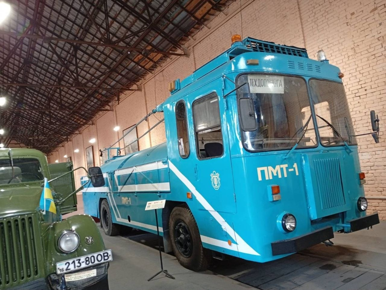 На світлині крупним планом зображено унікальний музейний експонат — спеціалізовану блакитну машину техдопомоги ПМТ-1 в Музеї електротранспорту в Одесі. Масивний спецавтомобіль із білими горизонтальними смугами на кузові обладнаний кабіною, що формою нагадує тролейбусну, та має напис «ТЕХДОПОМОГА» за лобовим склом. Поруч на корпусі нанесено офіційний білий герб Одеси, а перед капотом встановлено інформаційну табличку на металевій стійці. Зліва на передньому плані видно частину іншого ретро-автомобіля темно-зеленого кольору з радянським номерним знаком «16 213-80 ОВ» та маленьким прапорцем України на капоті. Виставка розміщена всередині ангара з високою металевою стелею та світлою цегляною стіною на задньому плані.