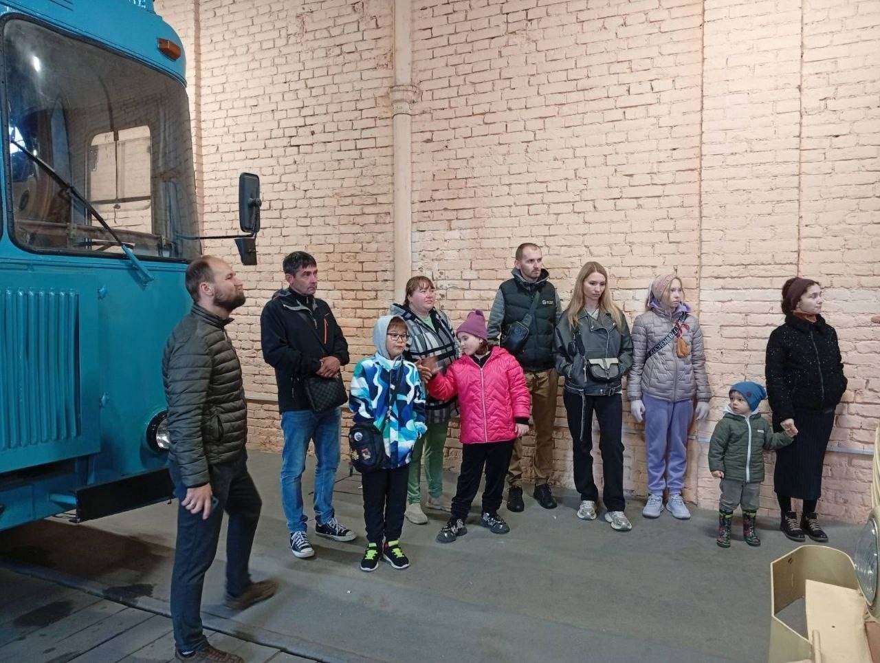 На світлині зображено родини внутрішньо переміщених осіб із Херсонщини під час екскурсії в Музеї електротранспорту в Одесі. Група з батьків та дітей стоїть на бетонній підлозі ангара на тлі високої світло-бежевої цегляної стіни та уважно слухає розповідь гіда. Ліворуч на передньому плані боком до об'єктиву стоїть чоловік у коричневій дутій куртці, а поруч із ним — інший чоловік у синіх джинсах і темній вітрівці. Серед дітей виділяються хлопчик у яскравому синьо-білому пуховику та дівчинка у рожевій куртці й фіолетовій шапці, яка вказує рукою вбік. На правому краю кадру жінка в чорному пальті тримає за руку найменшого хлопчика в синій шапці та зелених чобітках. Ліворуч у кадрі помітно передню частину масивного блакитного спецавтомобіля техдопомоги.