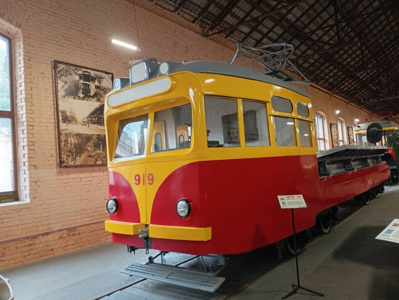 На світлині великим планом зображено унікальний історичний експонат — ретро-трамвай червоно-жовтого кольору з бортовим номером 919 в Музеї електротранспорту в Одесі. Передня частина вагону пофарбована у яскраво-жовтий та червоний кольори, має дві круглі фари, а на даху встановлено сіре комутаційне обладнання та штанговий струмоприймач. Поруч із вагоном на підлозі ангара виставлено інформаційну табличку на металевій стійці з написом «МТВ-82». На задньому плані вздовж світлої цегляної стіни з високими арочними вікнами розміщено велику вертикальну раму зі старовинними чорно-білими архівними фотографіями Одеси. Виставка облаштована всередині просторого екскурсійного депо з високою металевою стелею та рейками на підлозі.