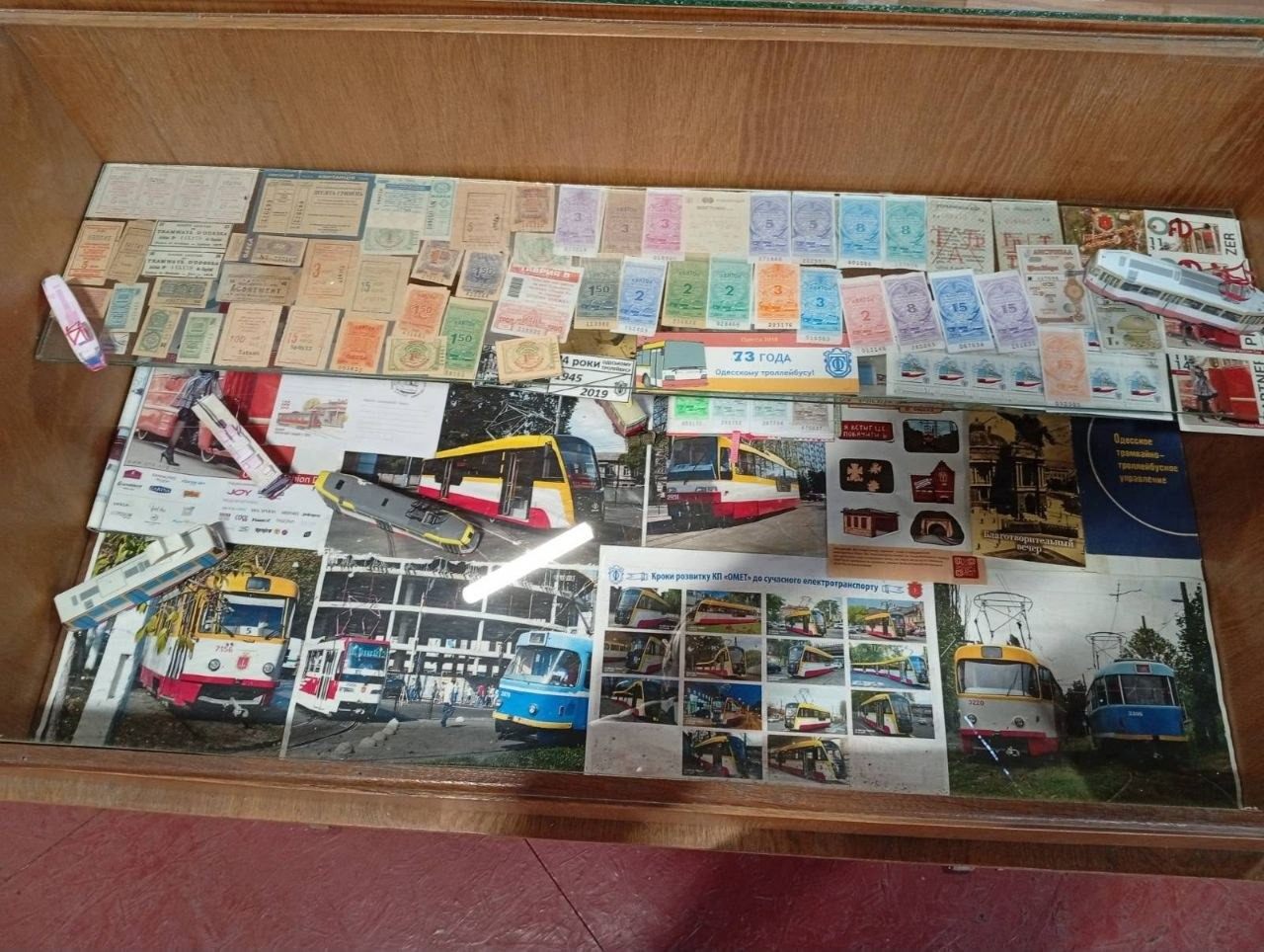 На світлині зафіксовано музейну скляну вітрину з унікальними експонатами в Музеї електротранспорту в Одесі. У верхній частині експозиції в кілька рядів розкладено велику колекцію старовинних різнокольорових паперових квитків та абонементів на проїзд у трамваях і тролейбусах різних років. Нижче під склом розміщені кольорові листівки, фотографії сучасних одеських трамваїв, інформаційний буклет «73 ГОДА Одесскому троллейбусу», брошура про кроки розвитку КП «ОМЕТ» та маленькі паперові макети моделей міського транспорту. Праворуч у вітрині лежить темна книга з золотистим написом «Одесское трамвайно-троллейбусное управление». Вітрина має дерев'яне обрамлення, а на склі видно світлові відблиски від ламп стелі.