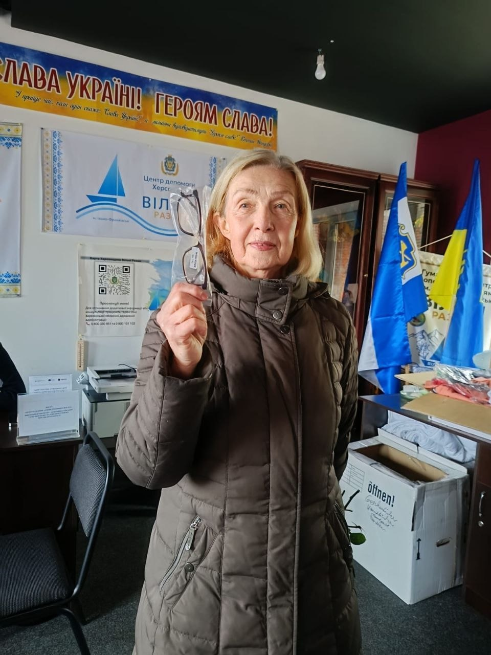 На світлині зображено жінку старшого віку з числа внутрішньо переміщених осіб з Херсонщини, яка отримала окуляри для корекції зору в координаційному центрі «Вільні разом» в Івано-Франківську. Вона стоїть по центру кадру в повен зріст у коричневому дутому пальті й тримає в піднятій правій руці нові окуляри у прозорому пластиковому пакуванні. Приміщення хабу облаштоване офісними меблями, великими картонними коробками гуманітарної допомоги з написами німецькою мовою та робочим місцем волонтерів ліворуч. На задньому плані кімнати на світлій стіні закріплено великий синьо-жовтий банер «СЛАВА УКРАЇНІ! ГЕРОЯМ СЛАВА!», брендоване полотно гуманітарного центру, а праворуч на флагштоках встановлено прапори, серед яких синій прапор Херсонської області та Державний Прапор України з патріотичними написами.