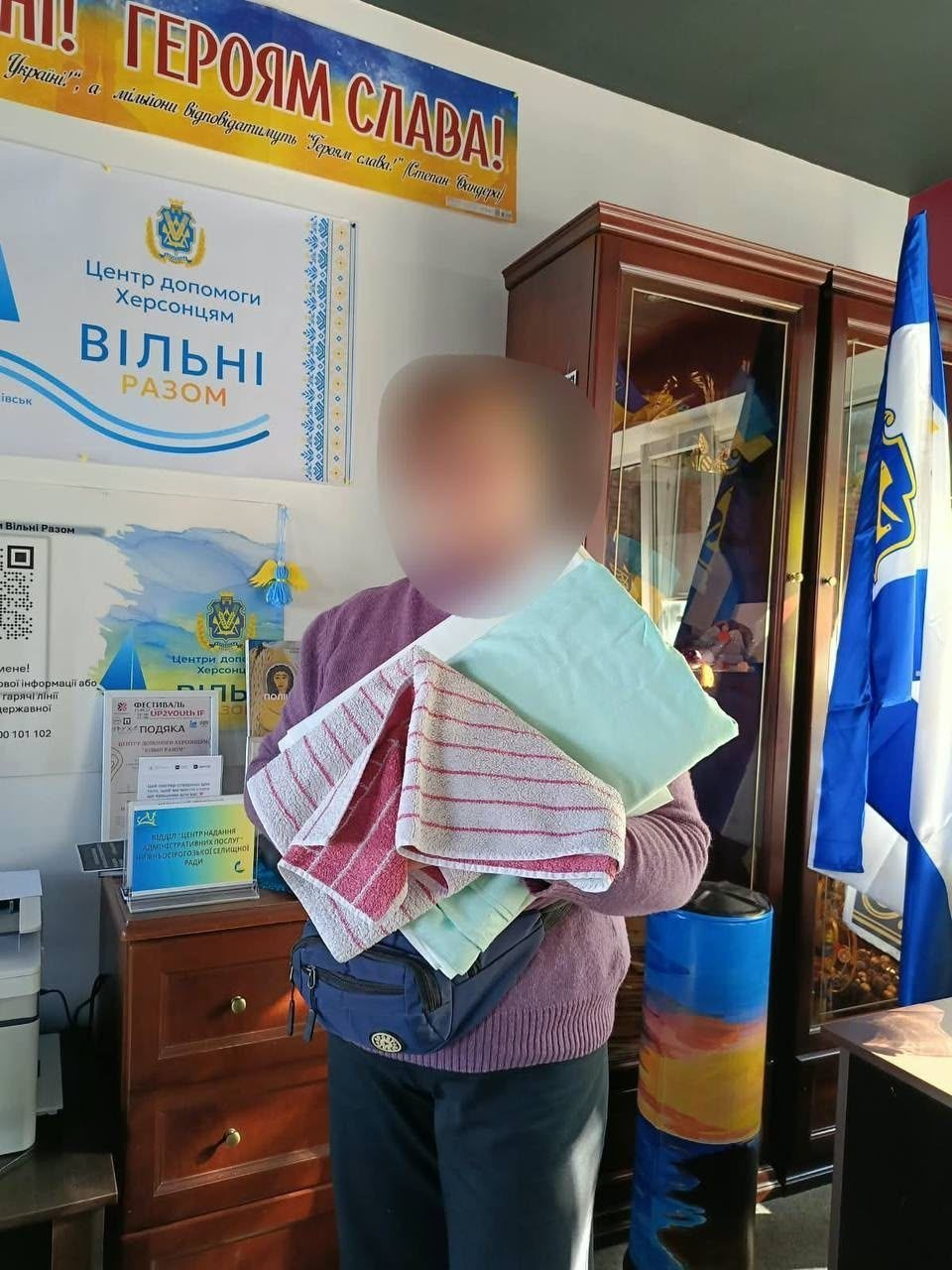 На світлині зображено жінку з числа внутрішньо переміщених осіб з Херсонщини, яка отримала домашній текстиль у координаційному центрі «Вільні разом» в Івано-Франківську. Вона стоїть по центру кадру, тримаючи в руках акуратно складений комплект нової постільної білизни світло-зеленого кольору та кілька різнокольорових рушників (зокрема рушник у біло-бордову смужку та зелений рушник). Переселенка одягнена у фіолетовий светр, темні штані та має через плече чорну сумку-бананку. На задньому плані кімнати хабу помітно масивну дерев'яну шафу-вітрину, брендований банер центру із написом «ВІЛЬНІ РАЗОМ», частину яскравого патріотичного плаката «ГЕРОЯМ СЛАВА!», а праворуч на флагштоку майорить синій прапор Херсонської області.