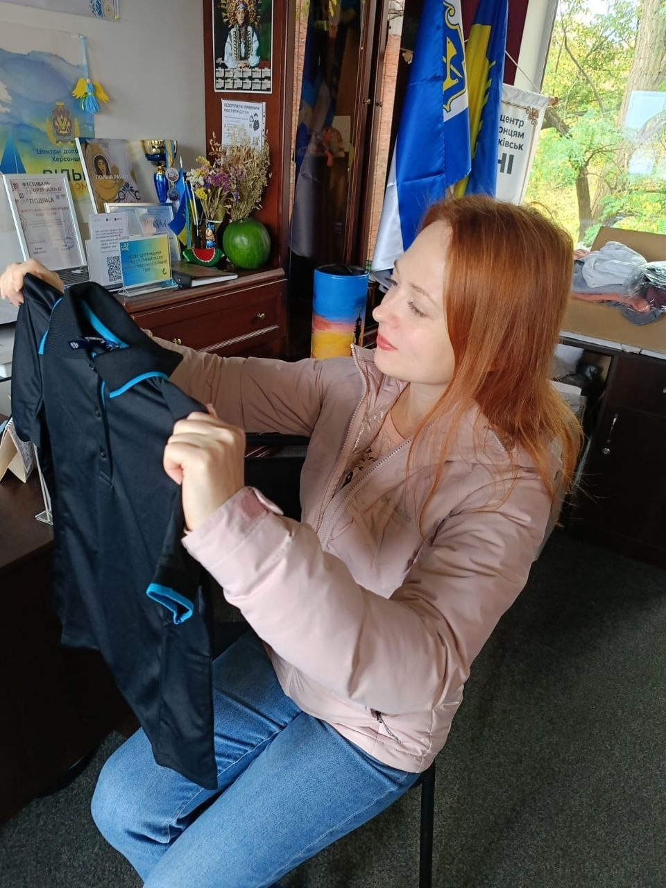 На світлині зображено момент видачі гуманітарної допомоги у координаційному центрі «Вільні разом» в Івано-Франківську. На передньому плані сидить жінка у світло-рожевій куртці та синіх джинсах, яка уважно розглядає чорну спортивну сорочку-поло з яскраво-блакитною окантовкою на комірі та рукавах. Робоче місце облаштоване в офісному приміщенні хабу, де на задньому плані помітно дерев'яну шафу з патріотичною сувенірною продукцією, прапор Херсонської області та Державний Прапор України на флагштоках. У кутку кімнати біля вікна складено картонні коробки з пакунками речей та сезонного одягу для внутрішньо переміщених осіб.
