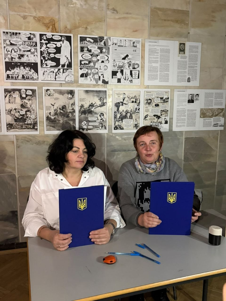 На світлині зображено момент підписання меморандуму про співпрацю між Департаментом реалізації гуманітарної політики Херсонської ОВА та ГО «Центр культурного розвитку «Тотем» з метою збереження спадщини художниці Поліни Райко. Ліворуч за сірим столом директор Департаменту реалізації гуманітарної політики Херсонської ОДА Віолета Царькова в білій блузі, а праворуч — представниця ГО «Тотем» у сірому світшоті з візерунком та кольоровому шийному шалику. Обидві тримають у руках сині офіційні папки з золотистим Державним Гербом України на тлі світлої кахельної стіни, яка завішена друкованими плакатами-мальописами виставки «Розпакована історія». На столі перед ними лежать дві сині ручки та канцелярська печатка.
