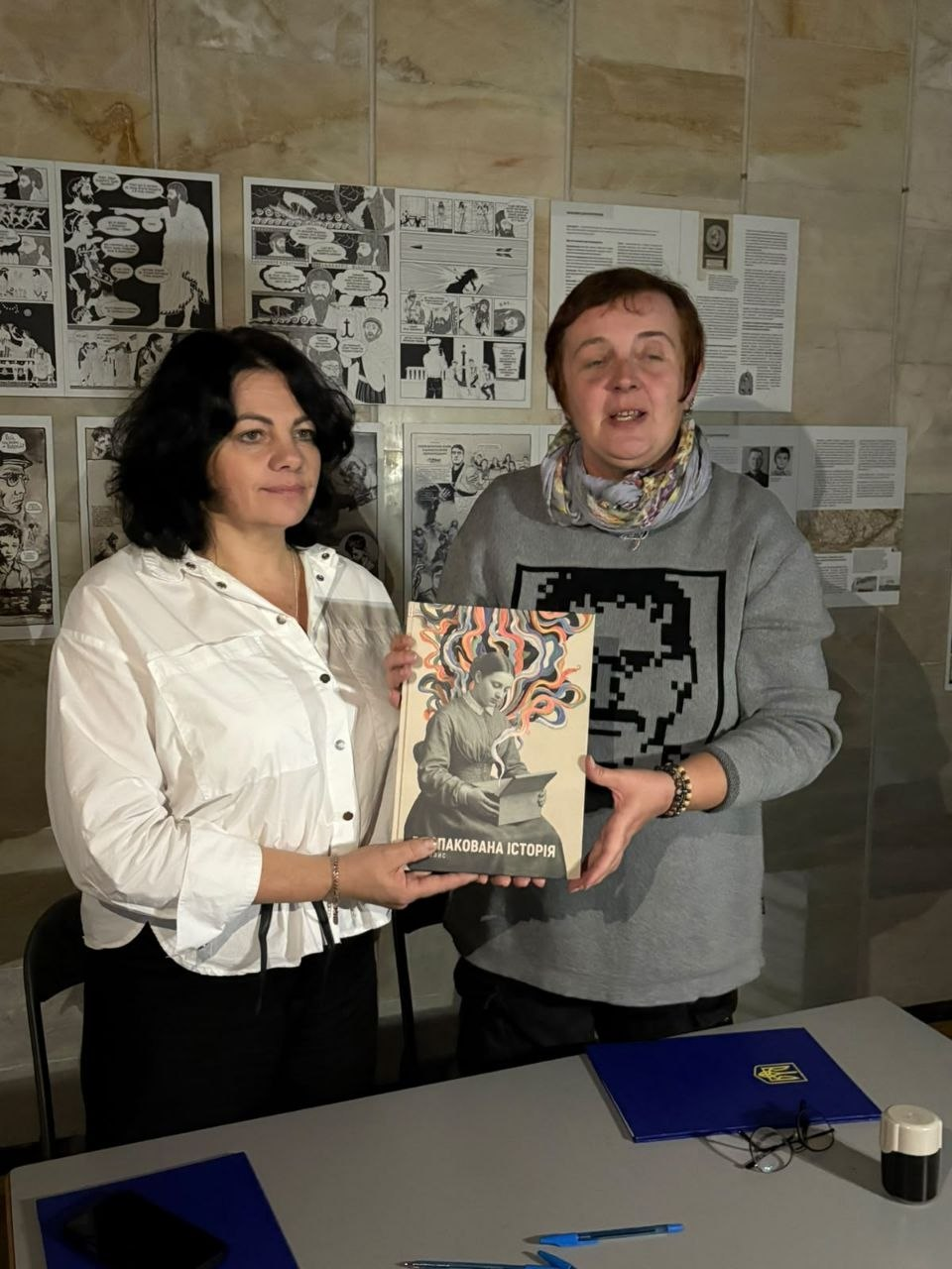 На світлині зафіксовано директора Департаменту реалізації гуманітарної політики Херсонської ОДА Віолету Царькову (ліворуч у білій блузі) та представницю ГО «Тотем» (праворуч у сірому світшоті), які разом тримають у руках велику книгу-мальопис із яскравою обкладинкою та написом «РОЗПАКОВАНА ІСТОРІЯ». Обидві жінки стоять біля сірого столу, на якому лежать синя папка, ручки, окуляри та печатка. На задньому плані кадру кахельна стіна зали завішена численними великими сторінками історичних коміксів та мальописів, що створюють єдину виставкову експозицію.