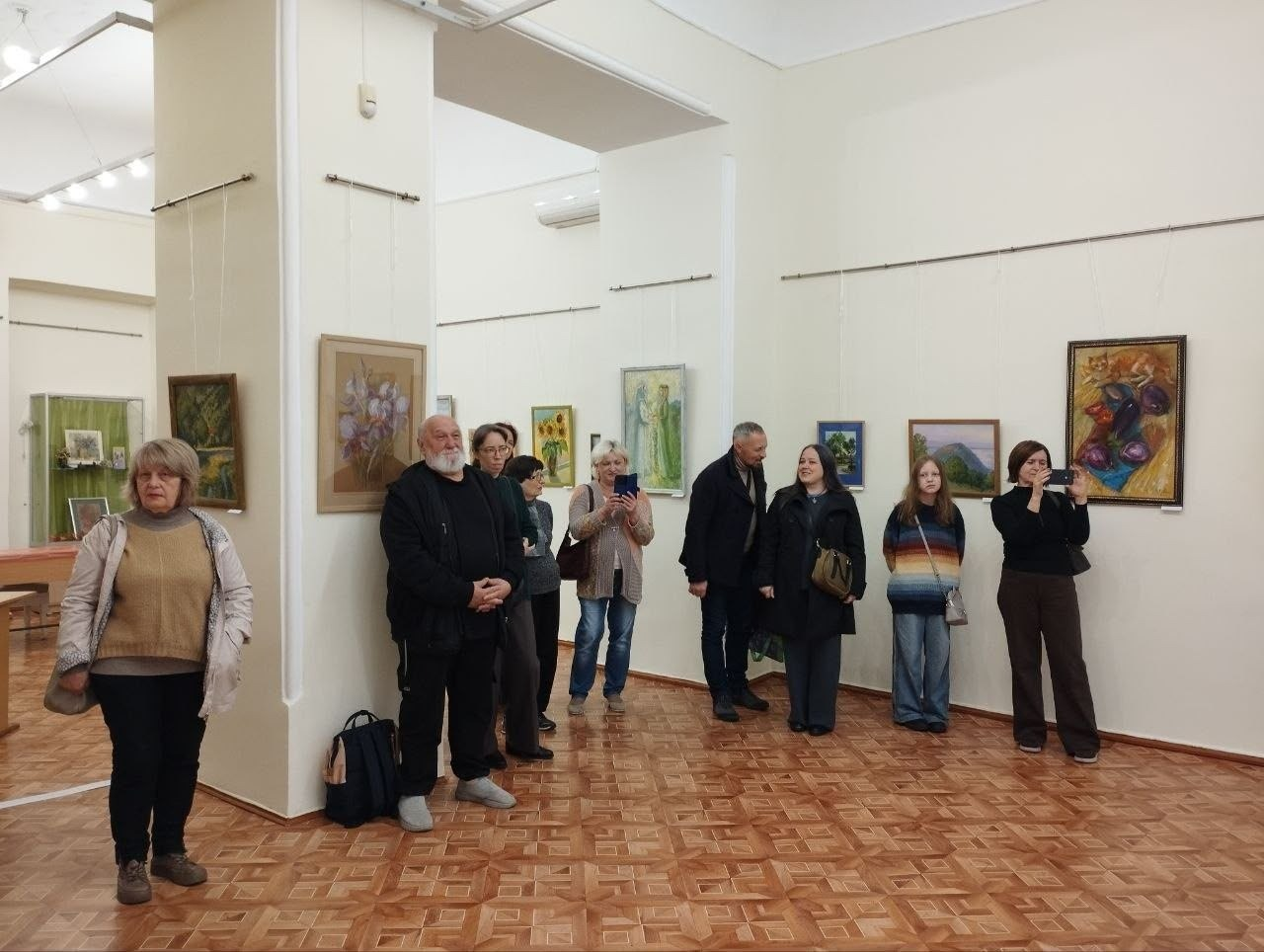 На світлині зображено внутрішньо переміщених осіб із Херсонщини в Одеському історико-краєзнавчому музеї під час огляду персональної виставки Ірини Рубель «Мої пори року». Відвідувачі розійшлися просторою виставковою залою з паркетною підлогою та світлими стінами, на яких розвішані різнокольорові картини художниці в рамах із зображенням пейзажів та натюрмортів. На передньому плані ліворуч стоїть жінка у світлій куртці та бежевому светрі, поруч із нею біля масивної колони позує сивочолий чоловік із бородою в чорному одязі, а інші гості розглядають експонати, дехто фотографує картини на смартфони. Захід організовано публічною бібліотекою Верхньорогачицької селищної ради.