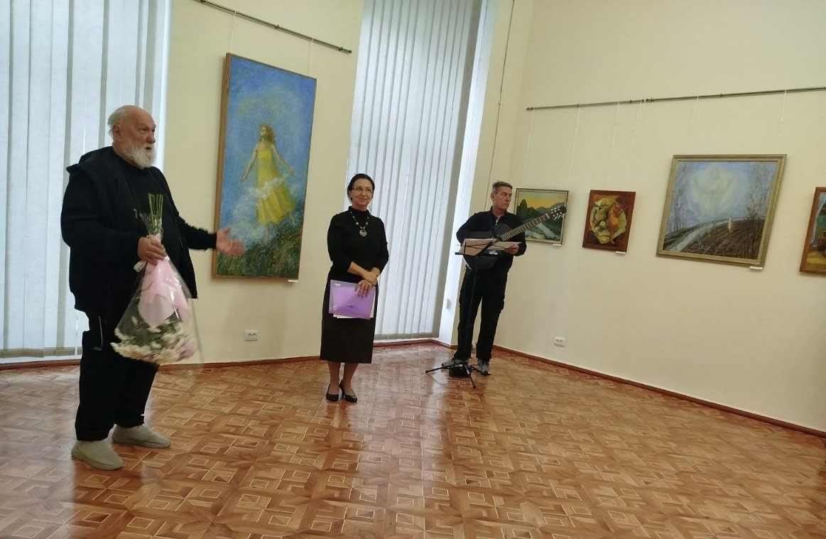 На світлині зафіксовано урочисту частину в Одеському історико-краєзнавчому музеї під час відвідування виставки переселенцями з Херсонщини. Ліворуч на передньому плані стоїть сивочолий чоловік із довгою білою бородою в чорному одязі, який емоційно виступає з промовою, тримаючи в правій руці великий букет білих квітів у рожевому пакуванні. Поруч із ним по центру стоїть жінка в елегантній чорній сукні з намистом та в окулярах, яка тримає перед собою фіолетову папку з документами й уважно слухає промовця. На задньому плані праворуч чоловік у чорному одязі акомпанує присутнім, граючи на акустичній гітарі біля пюпітра з нотами. Подія розгортається у просторій світлій залі з паркетною підлогою, великими вікнами із вертикальними жалюзі та розвішаними на стінах художніми картинами в рамах.