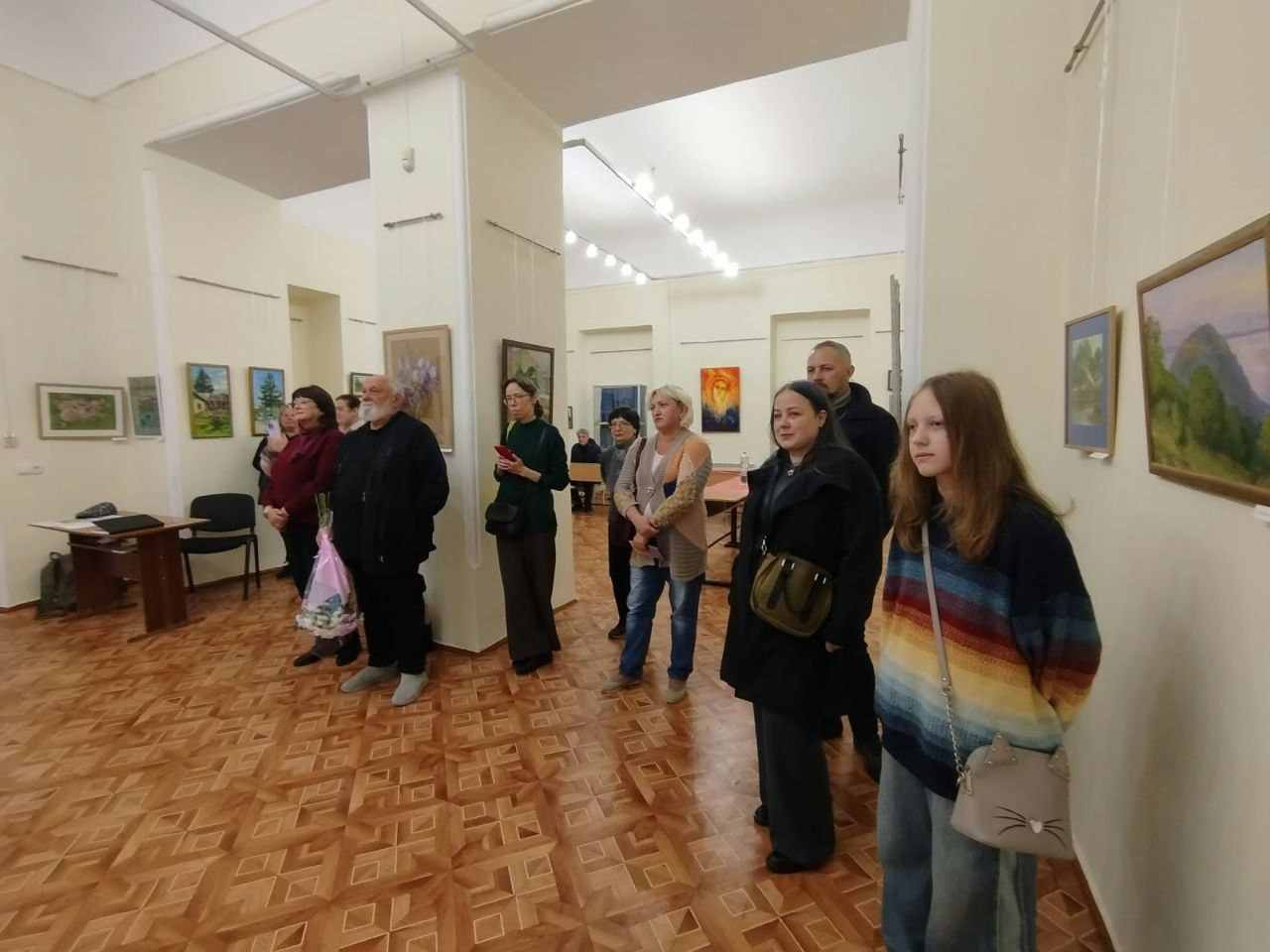 На світлині зафіксовано момент огляду виставки внутрішньо переміщеними особами з Херсонщини в Одеському історико-краєзнавчому музеї. Група відвідувачів зосереджена в лівій та центральній частинах зали, уважно слухаючи виступ або екскурсовода, який перебуває поза межами кадру. На передньому плані праворуч стоїть дівчинка-підліток у яскравому смугастому різнокольоровому светрі та з маленькою бежевою сумочкою з вушками через плече. Поруч із нею перебуває жінка в довгому чорному пальті з коричневою сумкою. У глибині зали біля колони стоїть знайомий сивочолий чоловік із білою бородою в чорному одязі, який тримає в руках отриманий раніше букет квітів у рожевому пакуванні. Світлі стіни приміщення та колони прикрашені живописними картинами в рамах, а на задньому плані помітно частину адміністративної зони зі столом та стільцями.