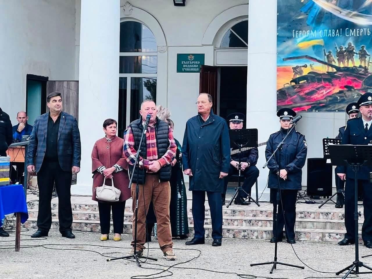 На світлині зафіксовано виступ начальника Чулаківської СВА Руслана Русаля з промовою біля мікрофона під час святкування Дня села Курісове. Очільник громади в картатій червоно-чорній сорочці та чорній дутій безрукавці стоїть по центру перед будівлею Будинку культури, де над входом розміщено зелену табличку болгарською мовою «БЪЛГАРСКО НЕДЕЛНО УЧИЛИЩЕ». Ліворуч за ним стоїть жінка в бордовому пальті з білою сумкою в руках, а крайнім ліворуч — чоловік у піджаку в клітинку. Праворуч від промовця перебуває чоловік у темно-синьому плащі та окулярах. На передньому плані праворуч стоять двоє співробітників поліції (чоловік та жінка) у форменому одязі біля стійок із мікрофонами, а позаду праворуч розмістився музичний ансамбль МВС. Права частина кадру прикрашена великим патріотичним банером із підбитою ворожою технікою та гаслом «ГЕРОЯМ СЛАВА! СМЕРТЬ ВОРОГАМ».