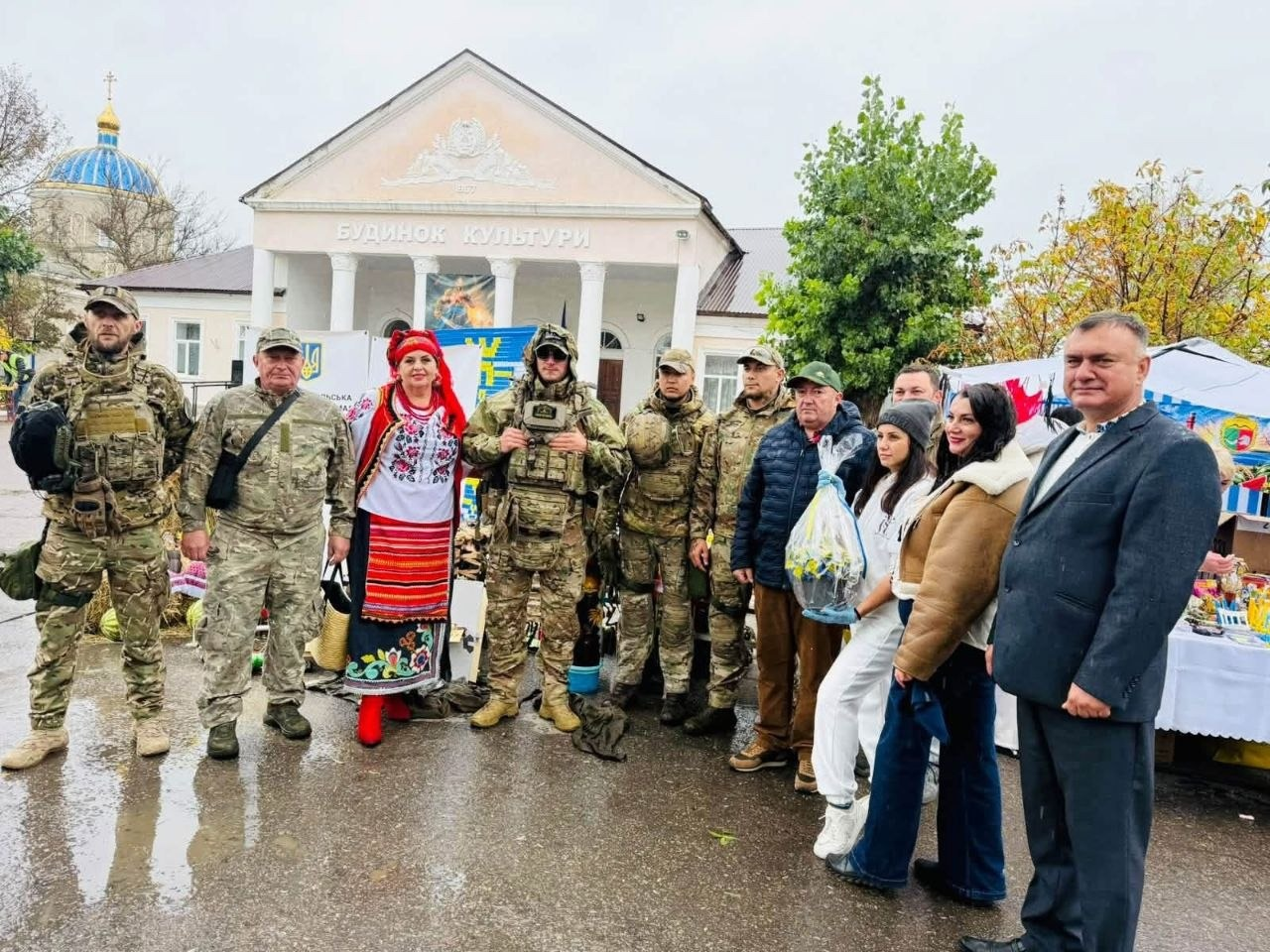 На світлині зафіксовано спільне фото делегації Херсонщини з українськими військовими та господарями свята на благодійному ярмарку в селі Курісове. Група з одинадцяти осіб вишикувалася в один ряд на вологому асфальті на тлі будівлі з написом «БУДИНОК КУЛЬТУРИ» та бані місцевого храму ліворуч. Серед присутніх виділяються четверо військовослужбовців ЗСУ у повному бойовому екіпіруванні та камуфляжі мультикам, жінка в українському національному костюмі з пишним вінком на голові, а також очільник Чулаківської СВА Руслан Русаль у зеленій кепці та темній куртці, який стоїть у центрі групи. Поруч із ним дівчина у білому костюмі тримає запакований у прозору плівку святковий коровай, а крайнім праворуч у кадрі стоїть чоловік у сірому класичному піджаку та білій сорочці.