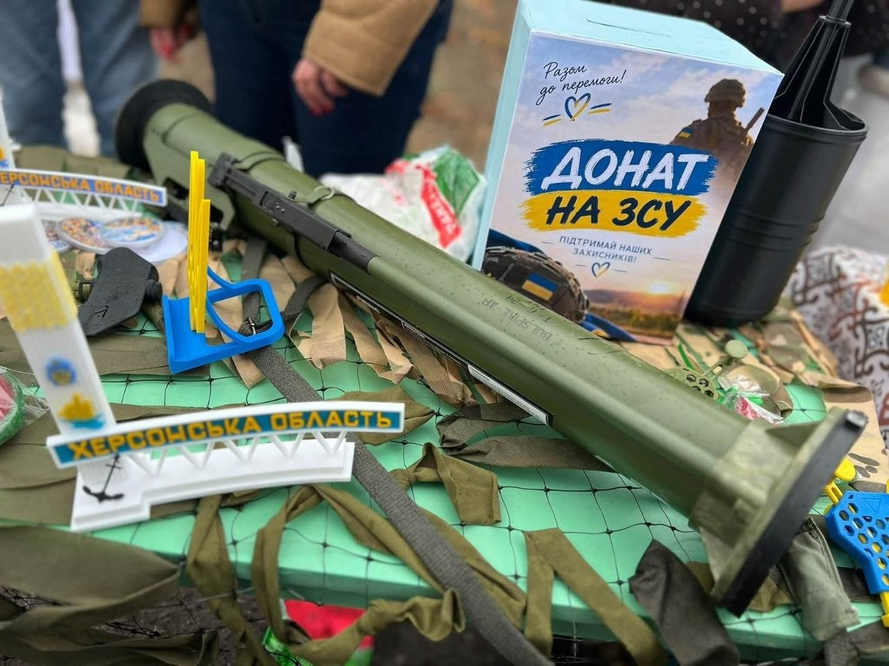 На світлині великим планом зображено експозицію благодійного ярмарку Чулаківської громади на підтримку Збройних Сил України. На столі, застеленому зеленою маскувальною сіткою, по діагоналі лежить зелений тубус від гранатомета із маркуванням. Праворуч стоїть блакитна картонна скринька для збору коштів із написом «ДОНАТ НА ЗСУ | Підтримай наших захисників!» та зображенням силуету військового. Ліворуч на передньому плані розміщено виготовлену на 3D-принтері мініатюрну білу копію знаменитої в'їзної стели з написом «ХЕРСОНСЬКА ОБЛАСТЬ», прикрашену Тризубом та синьо-жовтим прапорцем. Поруч розкладені інші дрібні патріотичні сувеніри та яскраві пластикові вироби хвостовиків для дронів, надруковані на 3D-принтері.